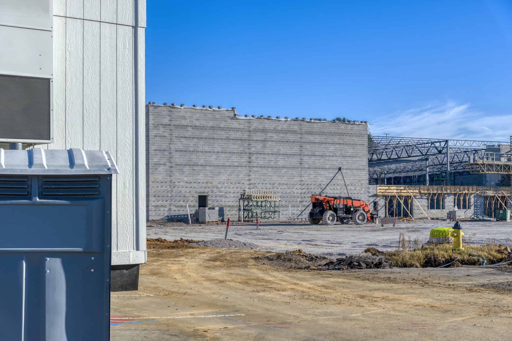 Orange telescopic forklift sits idle beside a half-built gray cinderblock wall, on a dirt construction site with scaffolding, concrete blocks, a yellow fire hydrant, and clear blue sky.