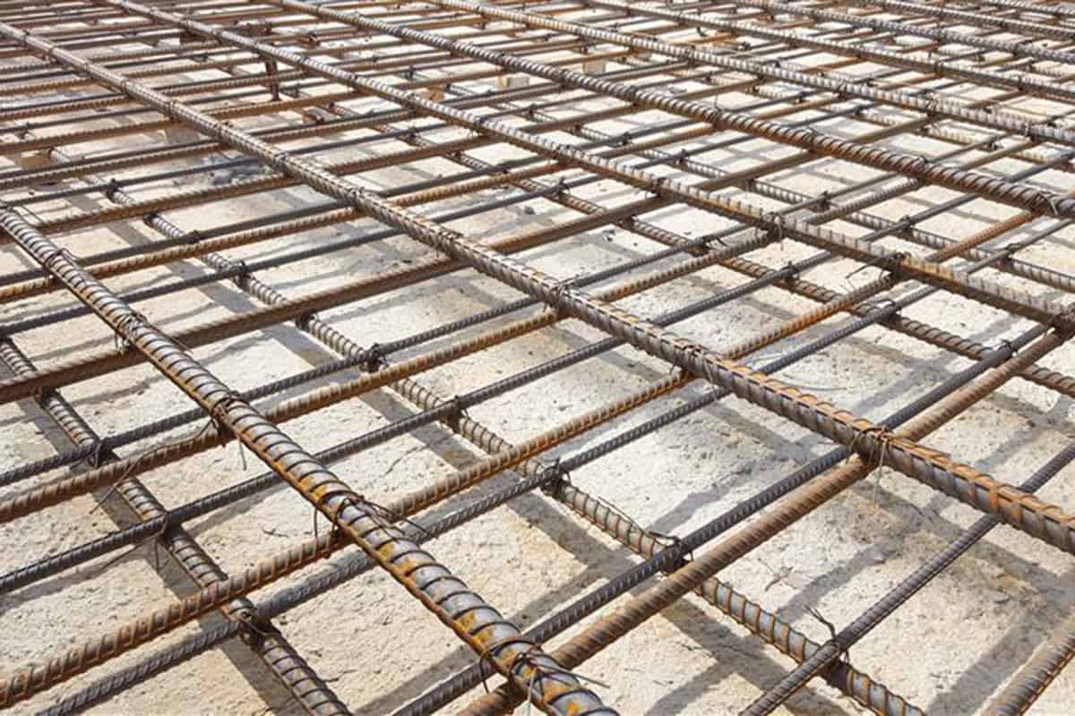 Steel rebar grid tied with wire forms a rectangular lattice over a sandy concrete slab at a sunlit construction site, casting strong parallel shadows.