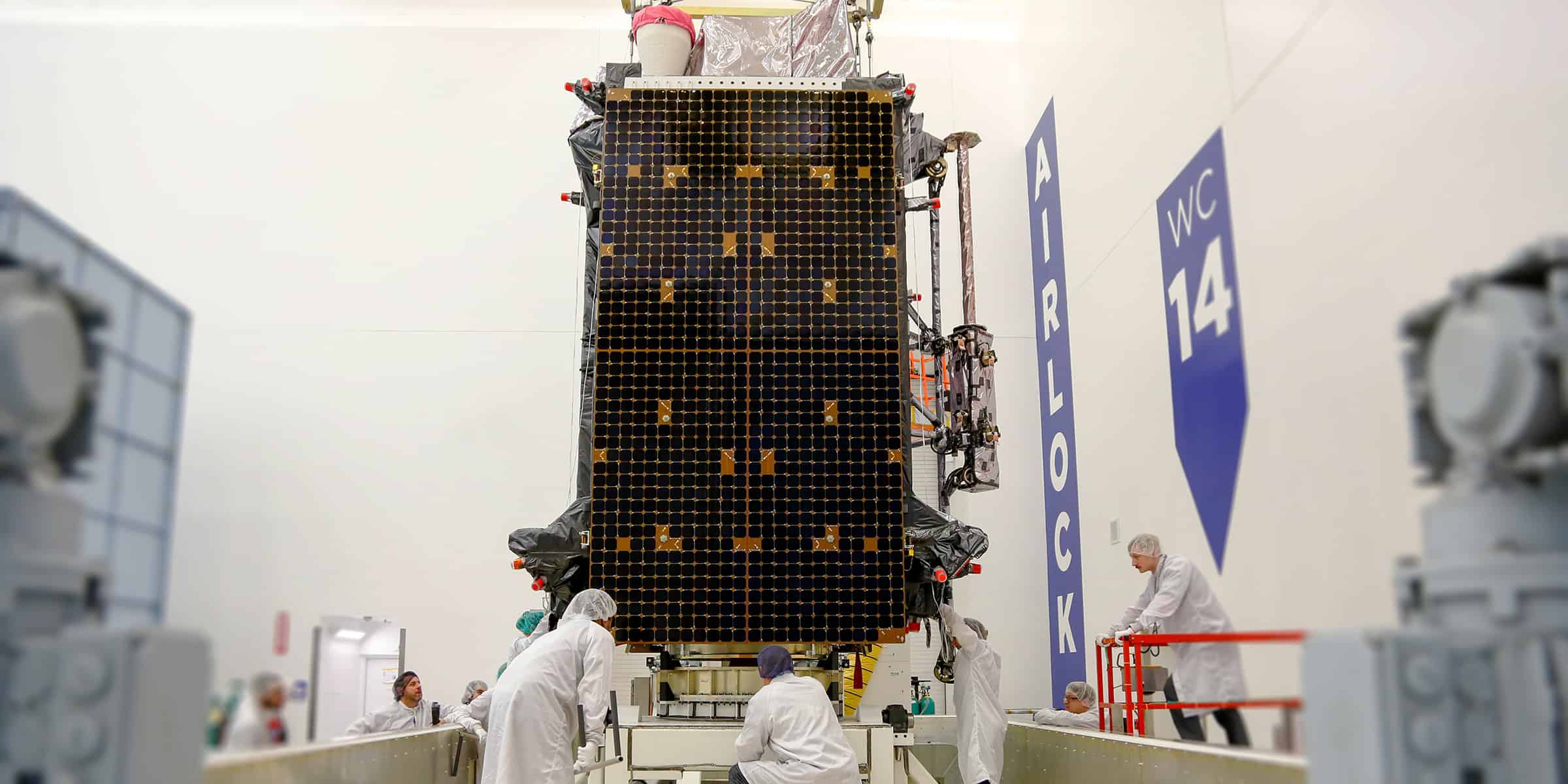 Large rectangular satellite solar-panel array — being lifted and aligned by technicians in white cleanroom suits — inside a white high‑ceiling integration room. Text in image: "AIRLOCK" and "WC 14".