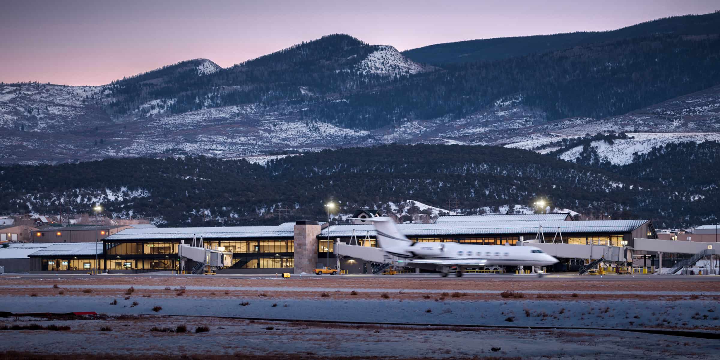 Private jet taxiing past a small, lit airport terminal at dusk; snow-dusted runway and low hills give way to forested, snow-capped mountains in the background.