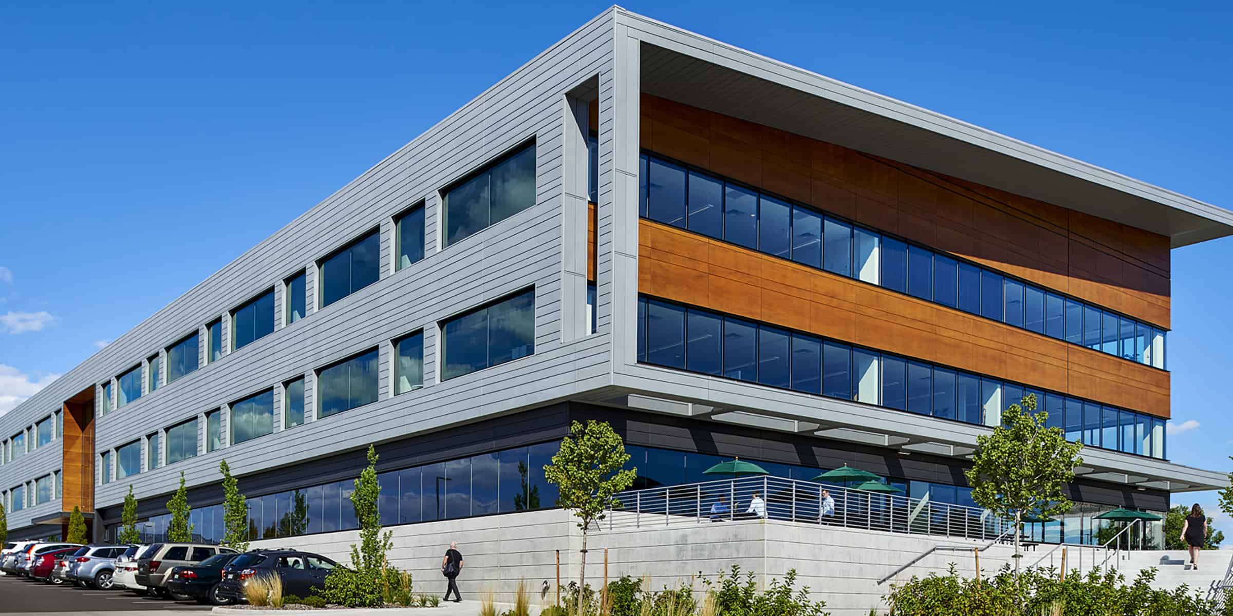 A modern four-story office building features large glass windows and wood-accented panels, projecting over a concrete terrace; cars parked, few people walk and sit under green umbrellas, clear blue sky.