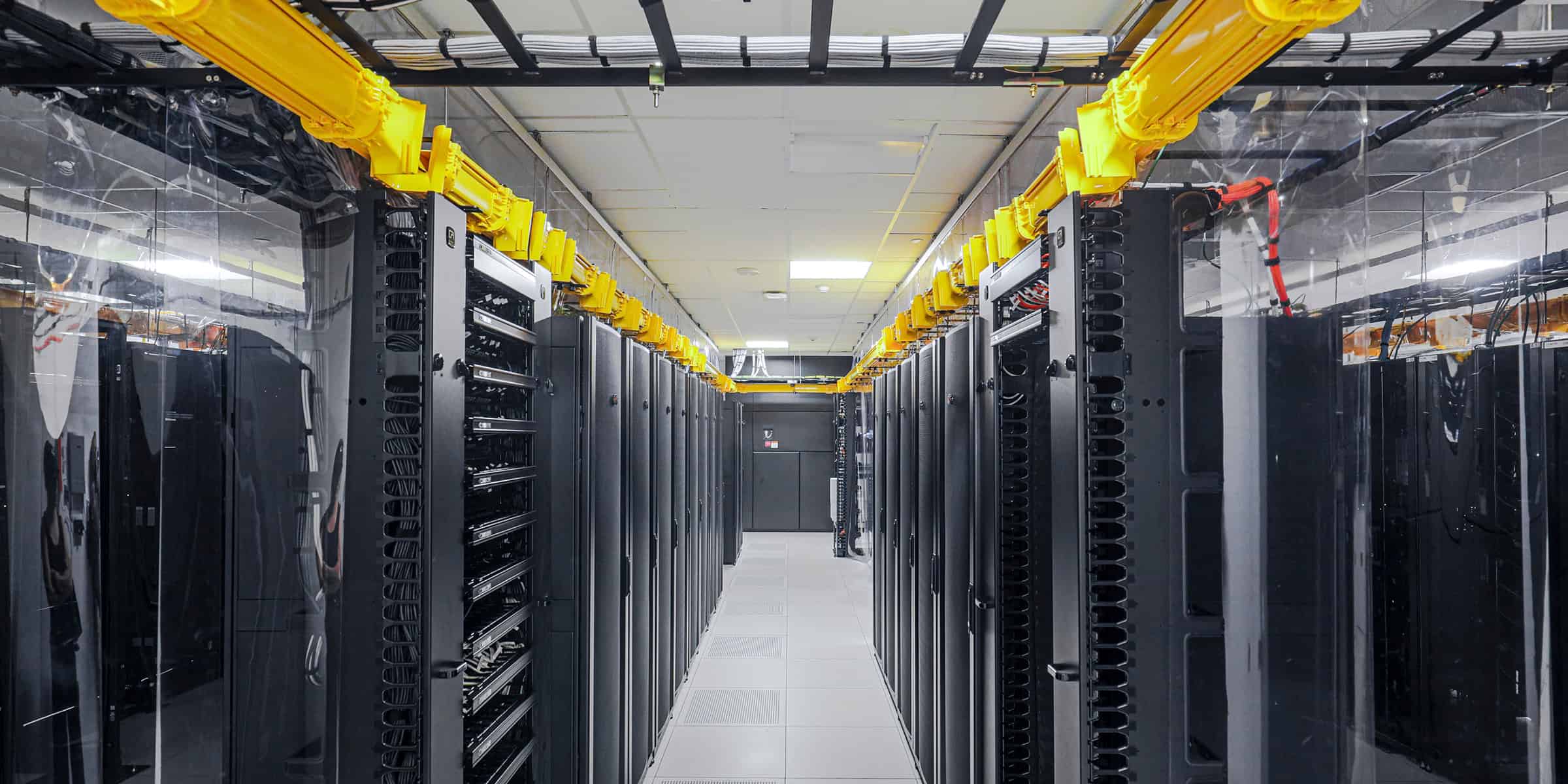 Server racks line a central aisle, housing network equipment interconnected by cables; bright yellow overhead cable trays run above, inside a clean, climate-controlled data center with raised floor tiles.