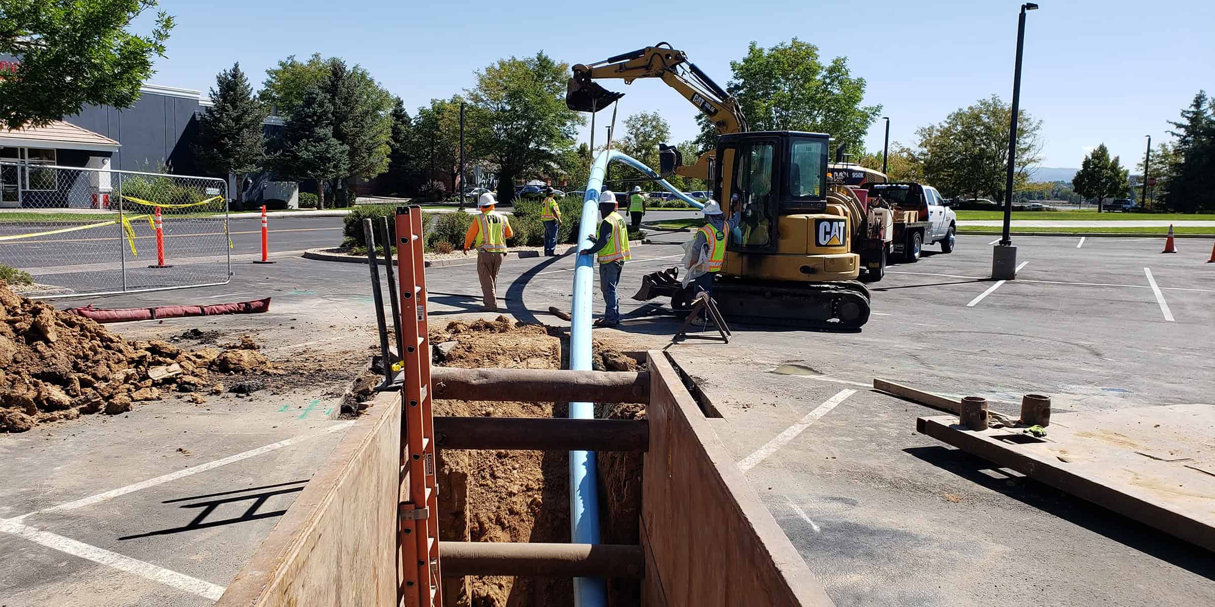 A light-blue pipe is being lowered into a trench; construction workers in safety vests guide it with an excavator amid dirt piles and cones in a sunlit parking lot by trees.

Text visible: "CAT" (on the excavator)