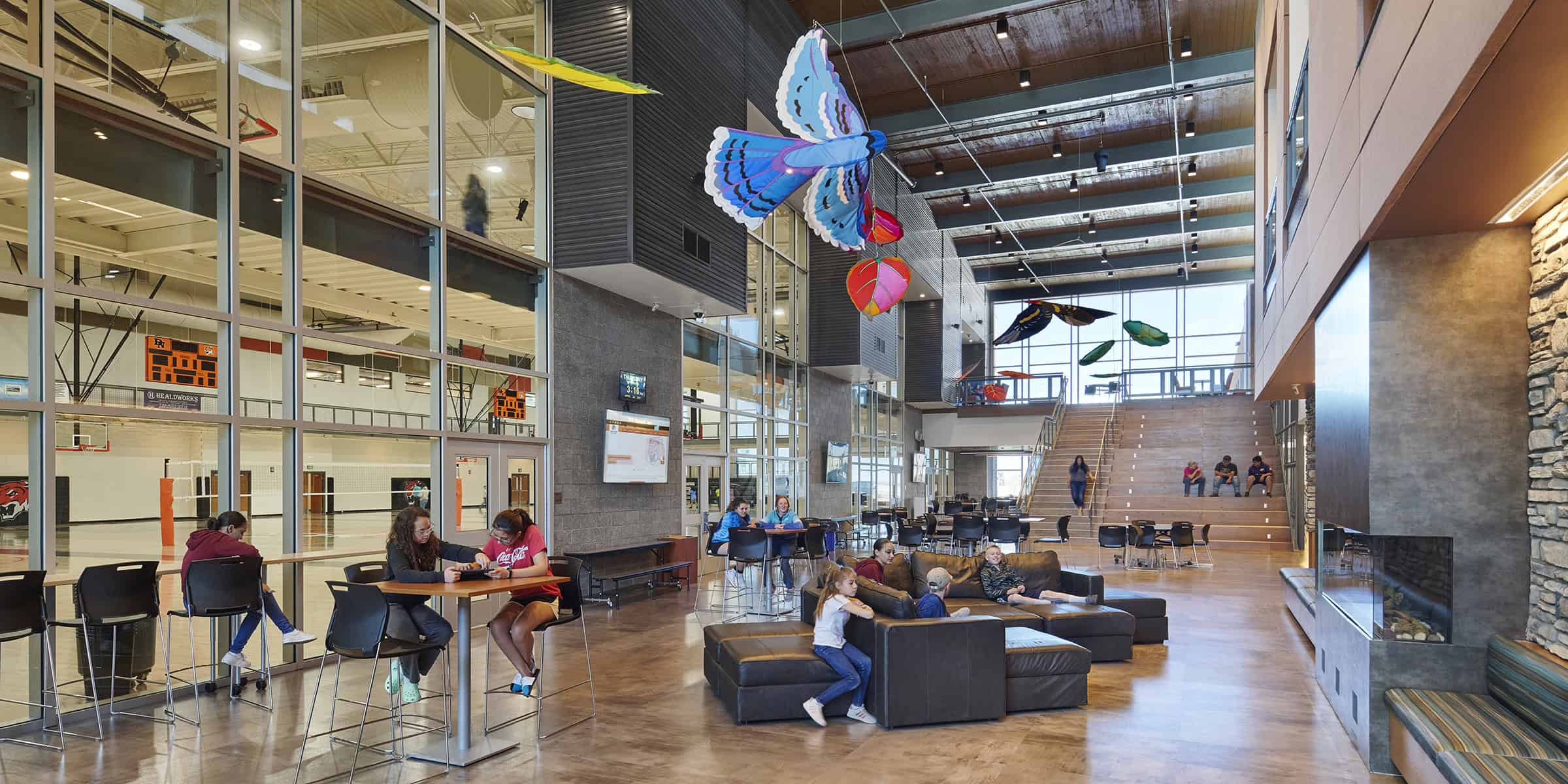 Large atrium seating area with leather sofas where people sit and talk; colorful butterfly kites hang overhead; glass wall reveals an indoor gym and tiered wooden stairs in a modern community center.

Text visible: "Coca-Cola" on a red shirt; digital clock reads "3:16".