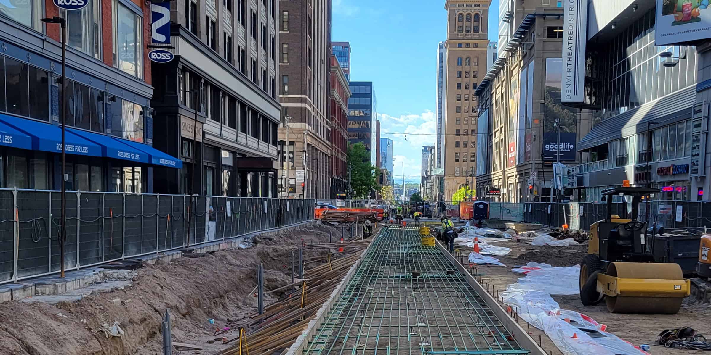 Long reinforced concrete slab (rebar) being laid by construction workers in a fenced downtown street, between tall commercial buildings under a clear blue sky.

Transcribed text: ROSS; ROSS DRESS FOR LESS; DENVERTHEATREDISTRICT; goodlife; CAT