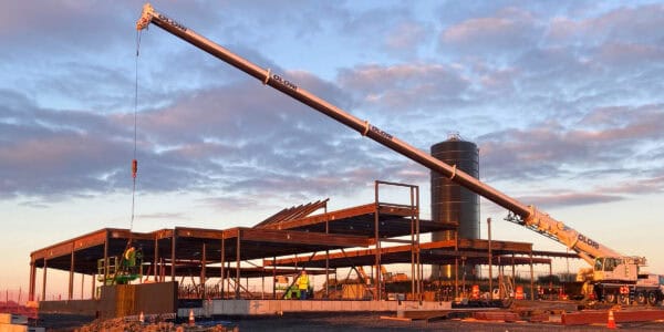 Large mobile crane extends its boom, lowering a hook over a partially built steel-frame structure amid a sunset construction site with a tall cylindrical silo and workers. Text: "OLORI" on boom.