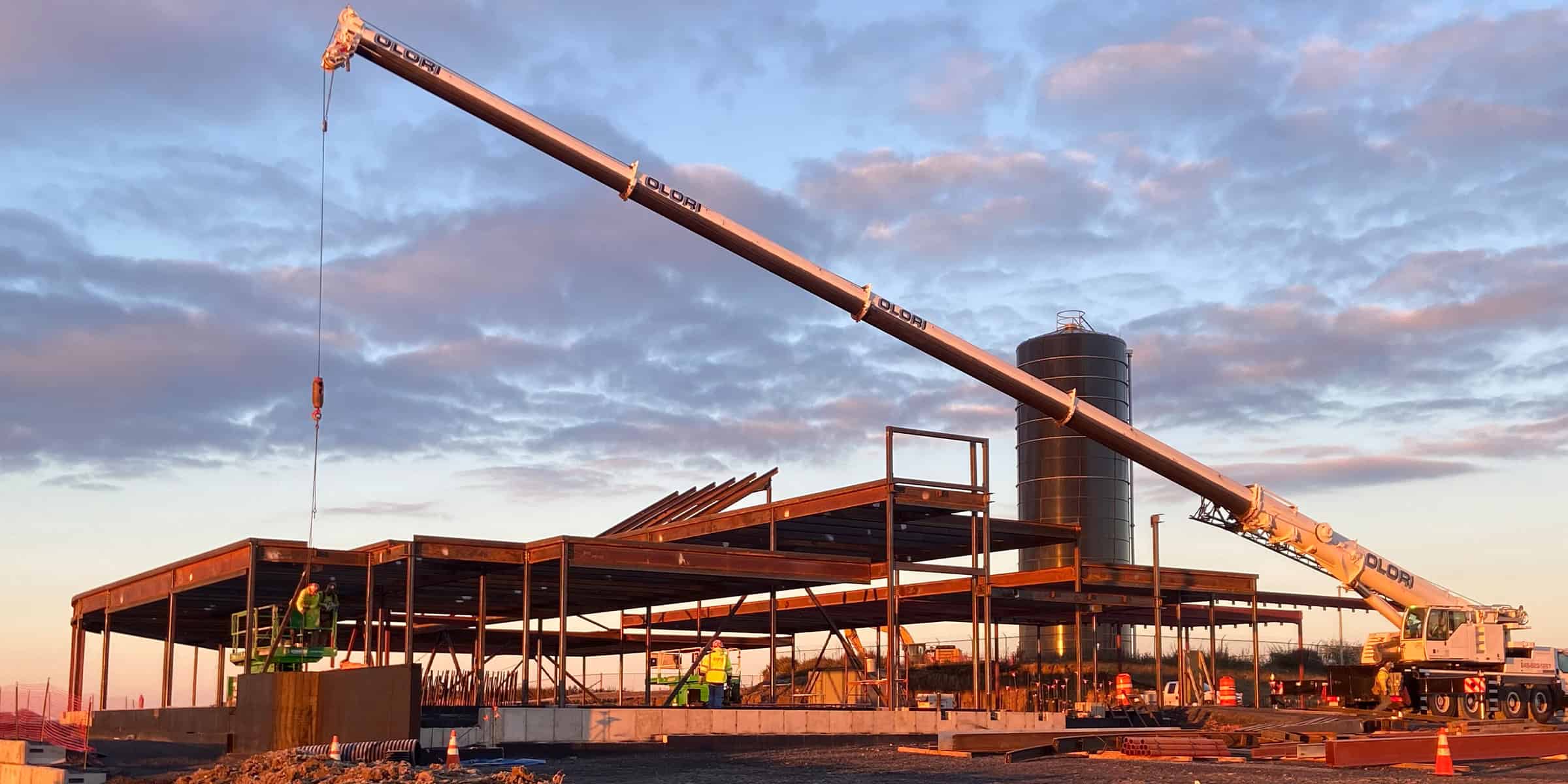 Large mobile crane extends its boom, lowering a hook over a partially built steel-frame structure amid a sunset construction site with a tall cylindrical silo and workers. Text: "OLORI" on boom.
