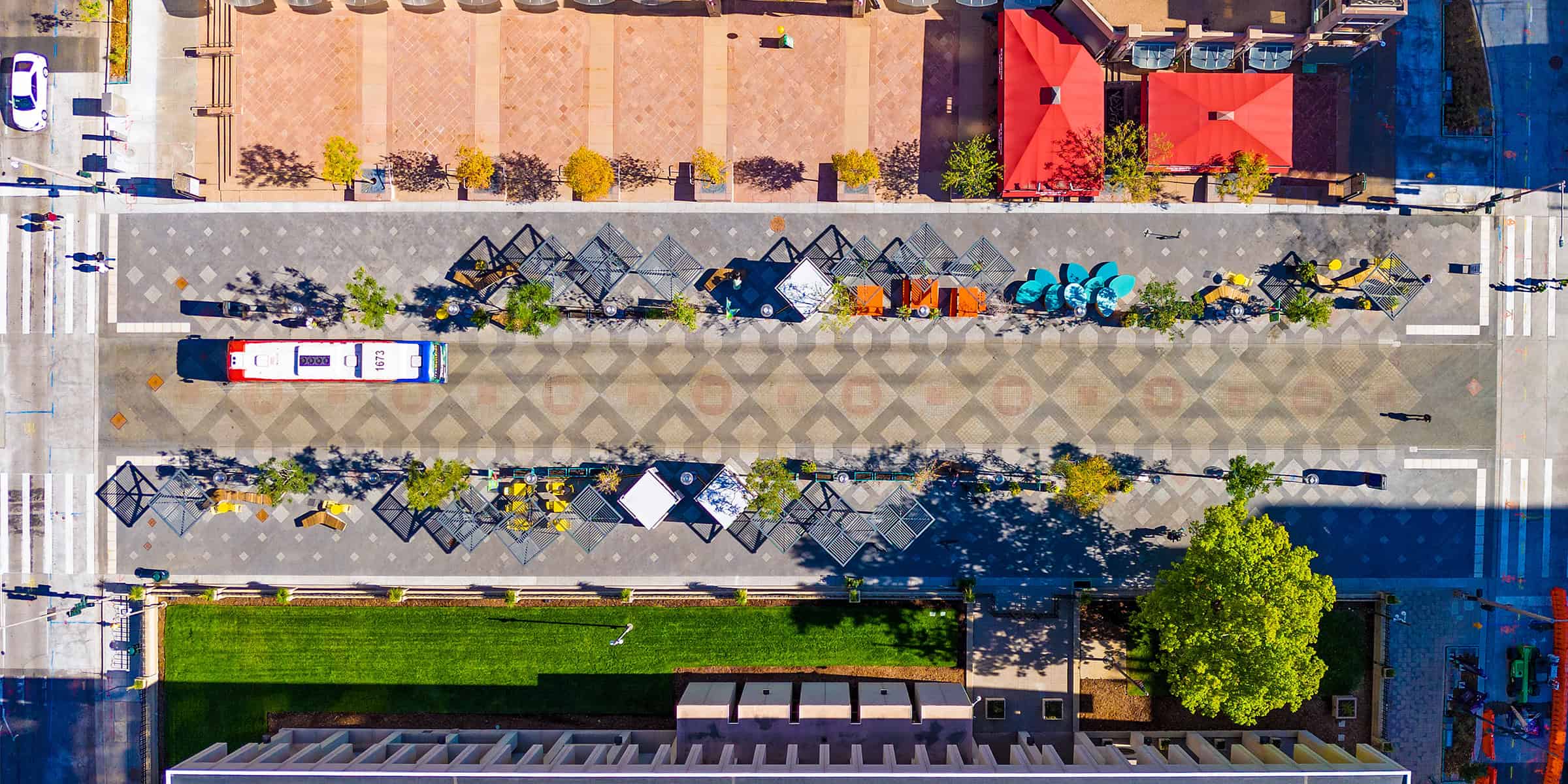 Bus number 1673 occupies a central paved median; surrounded by geometric tile patterns, shaded seating and trees, red-roofed buildings and a grassy lawn in an aerial urban plaza.