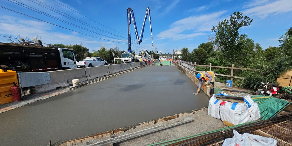 Fresh concrete slab being smoothed by a worker in a high-visibility vest and hard hat; concrete-pump trucks and crews operate on a partially closed roadway bridge amid trees and blue sky.