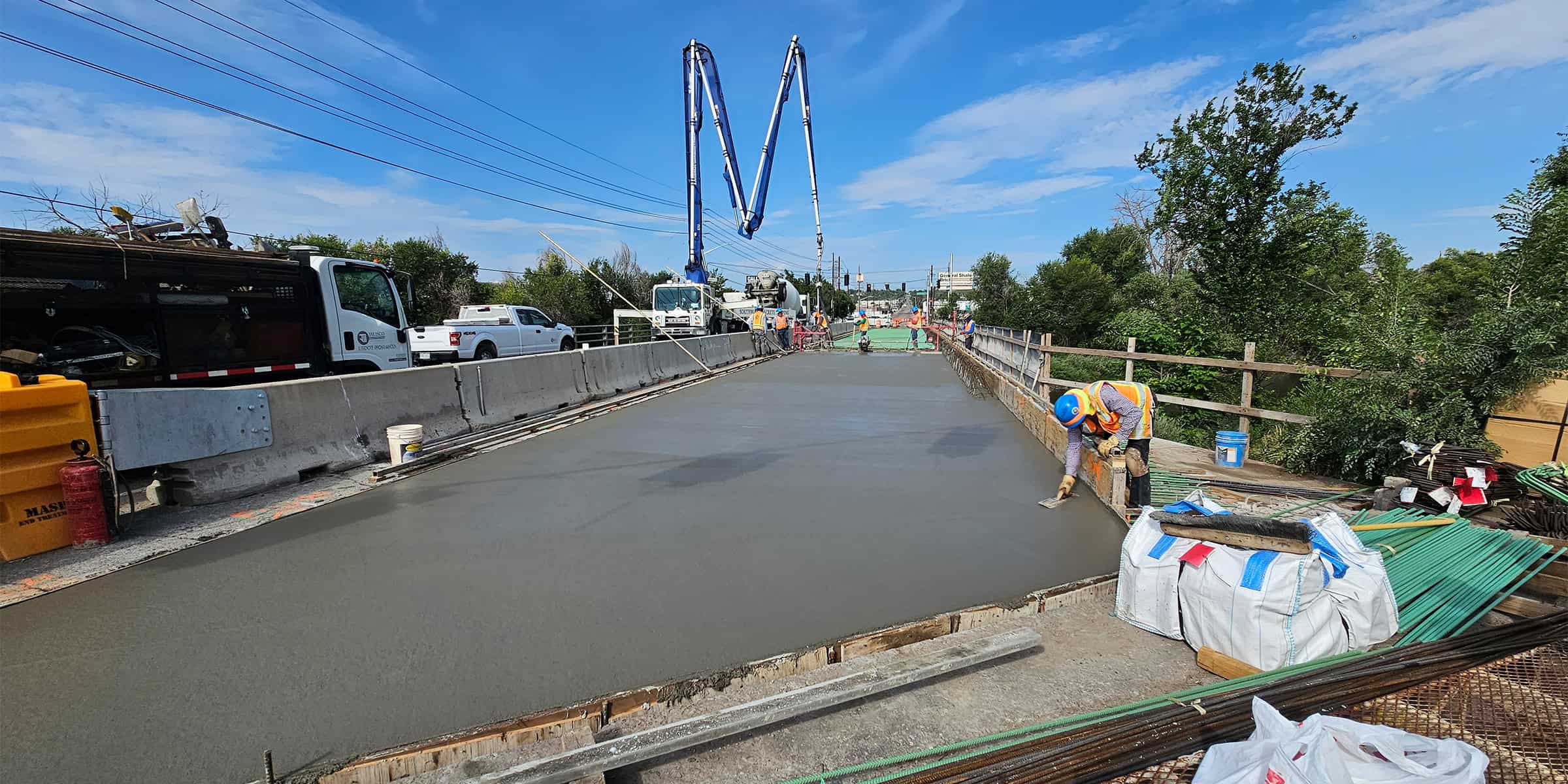 Fresh concrete slab being smoothed by a worker in a high-visibility vest and hard hat; concrete-pump trucks and crews operate on a partially closed roadway bridge amid trees and blue sky.