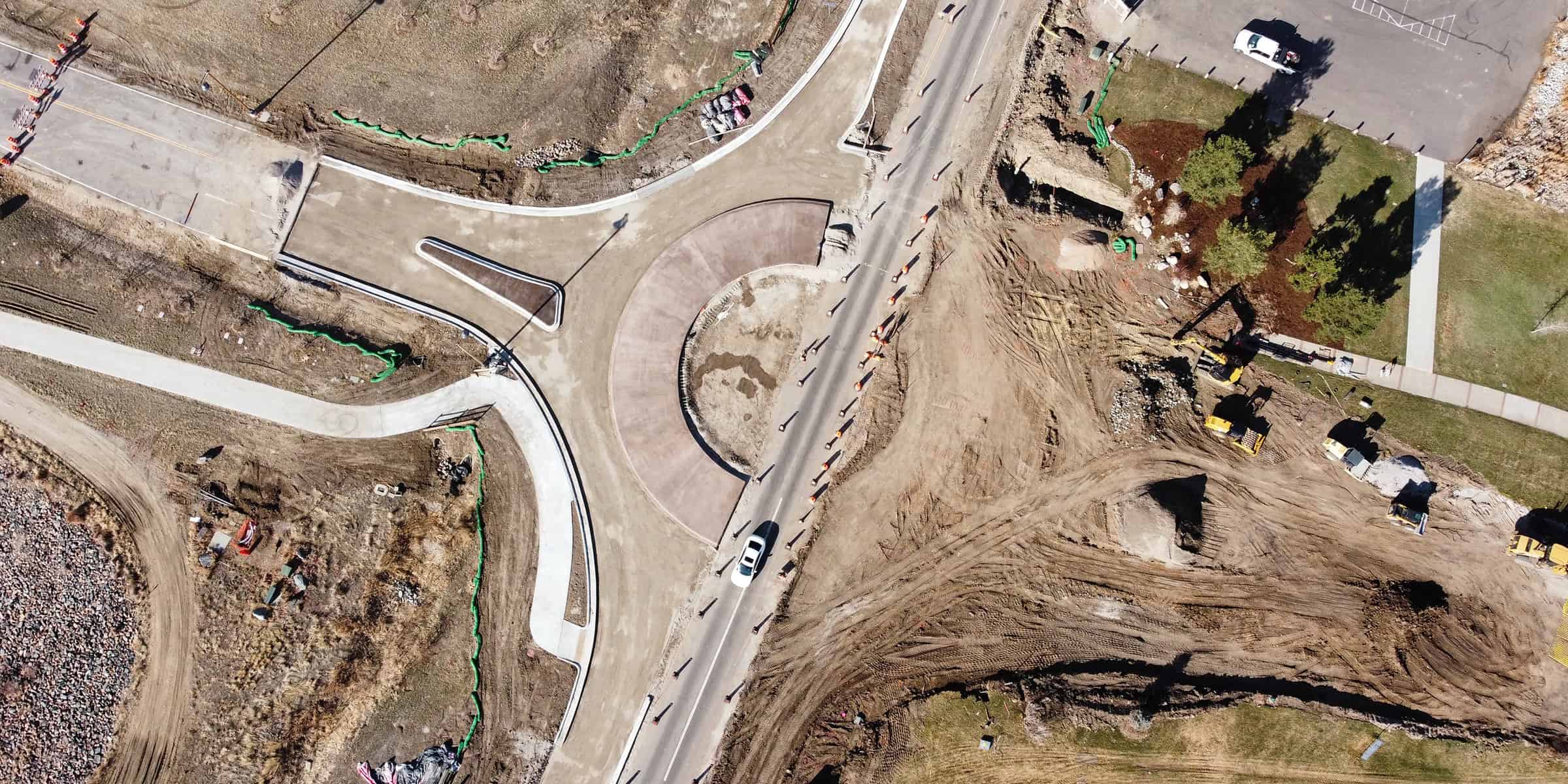 Roundabout is being built — partially paved center and curbs, with a white car passing; surrounded by dirt, construction equipment, orange cones, and nearby grassy sidewalks.