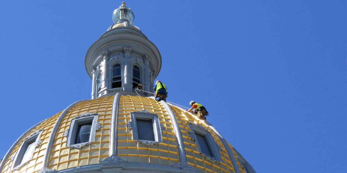 Gilded dome: two construction workers wearing hi‑vis vests and helmets climb and work on the curved gold‑tiled surface, secured by ropes, beneath a clear blue sky.