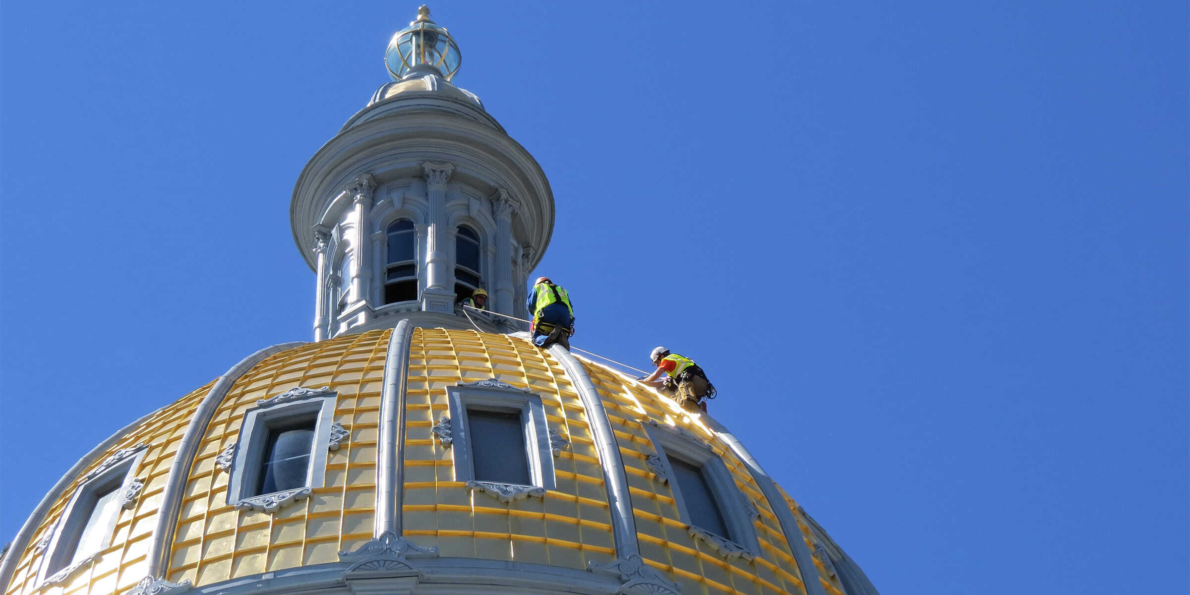 Gilded dome: two construction workers wearing hi‑vis vests and helmets climb and work on the curved gold‑tiled surface, secured by ropes, beneath a clear blue sky.