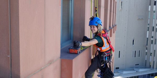 A female rope-access worker reaches to place an orange equipment box on a windowsill while suspended by ropes, wearing a blue helmet, orange vest and harness on a high-rise facade.