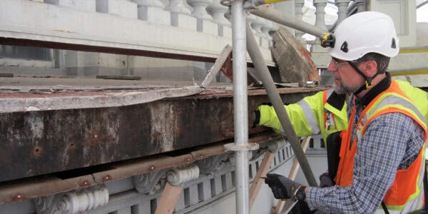 Corroded steel beam being inspected by a construction worker wearing a white hard hat, safety vest, and gloves while on scaffolding beside ornate stone balustrade and building facade.