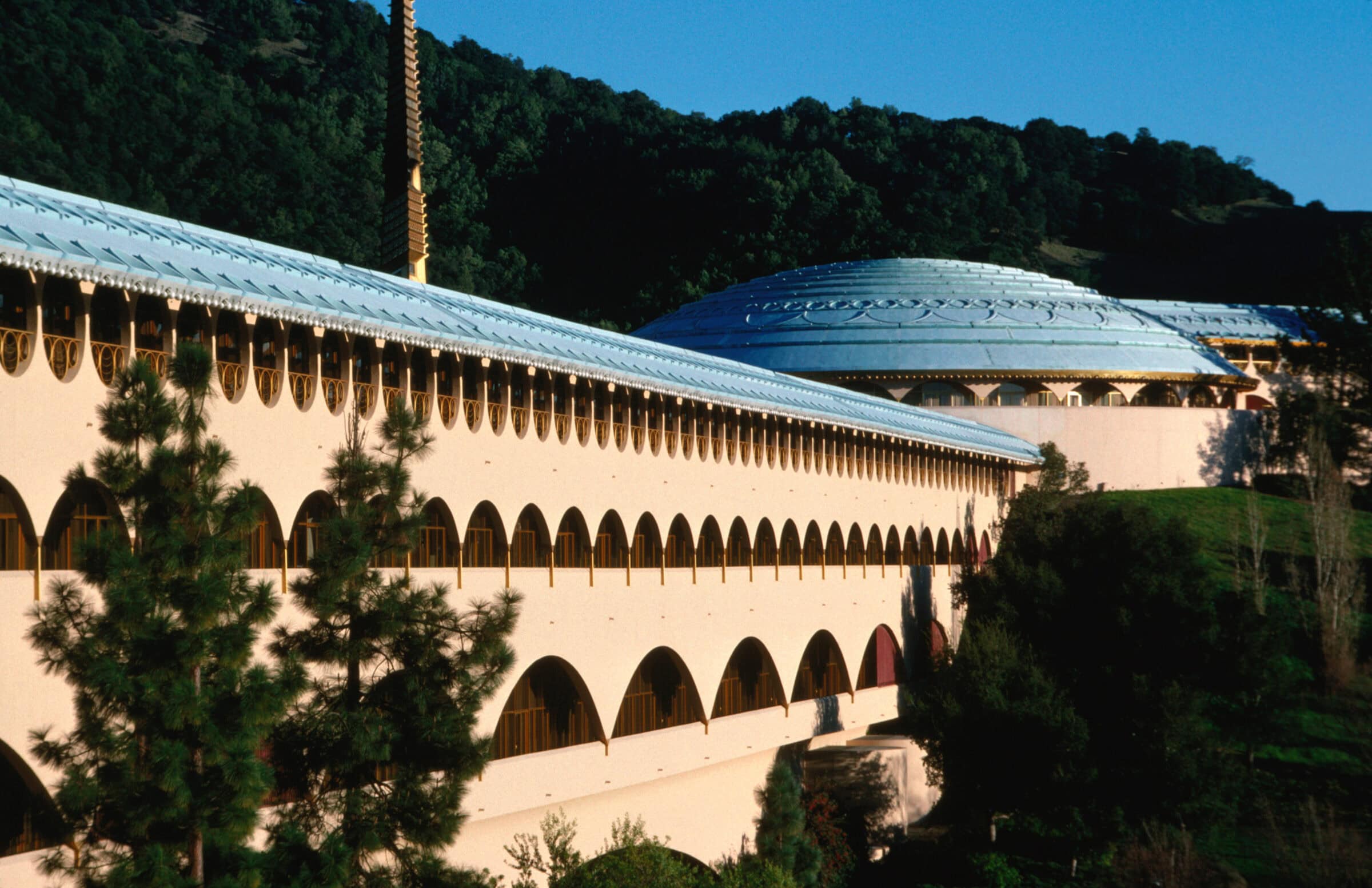 Long pale arched building stretches along a hillside, capped by a large patterned dome; evergreen trees sit in the foreground, with forested hills and clear blue sky beyond.