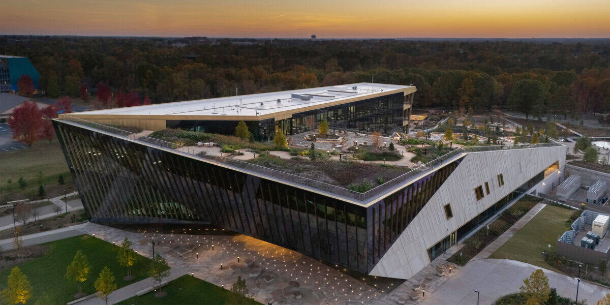 A triangular, modern building with a planted rooftop garden and glass facade features a landscaped terrace and illuminated entrance, set within wooded grounds and pathways at sunset.