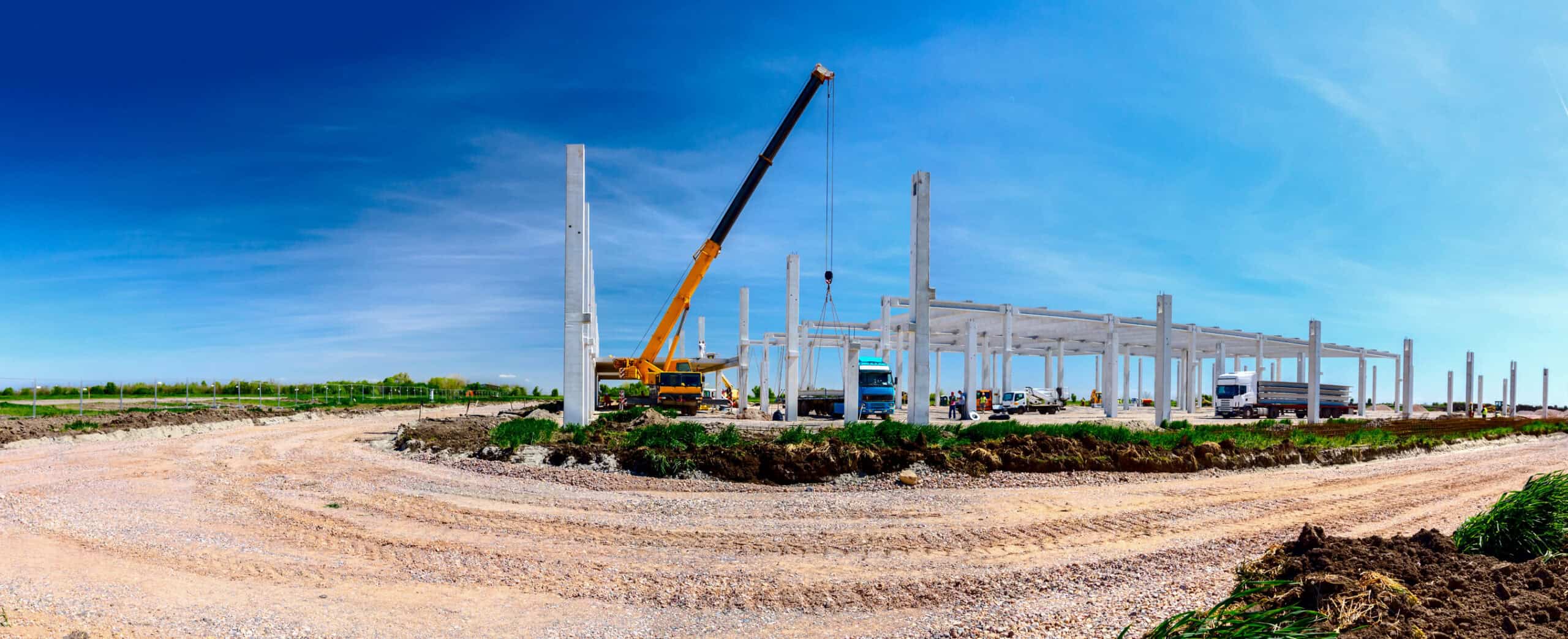A yellow crane lifts a concrete beam onto a skeletal concrete frame while trucks and workers operate around a gravel construction site beneath a clear blue sky.