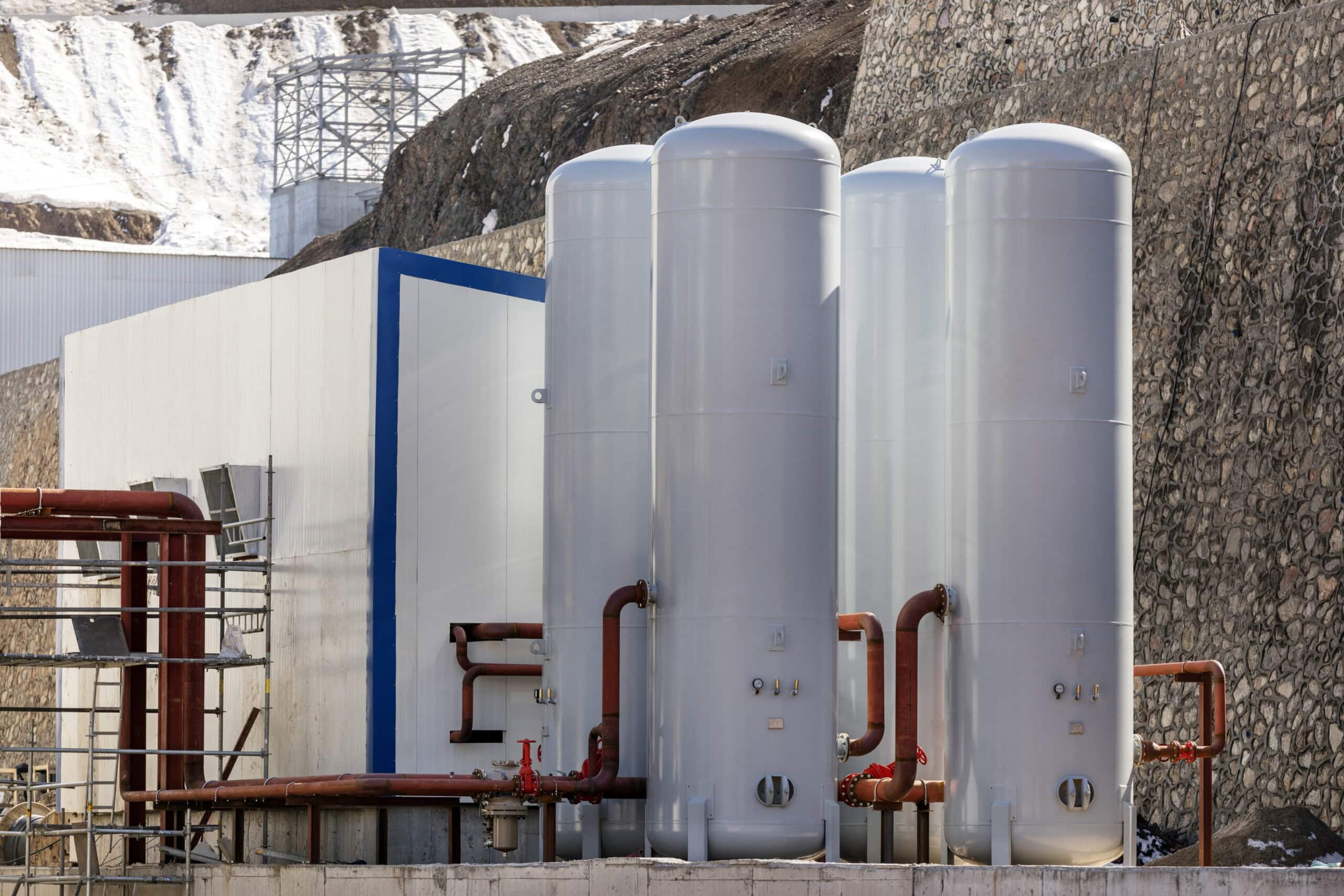 Four tall white cylindrical storage tanks stand connected by red piping and valves at an industrial facility, situated before a stone retaining wall and a snowy hillside.