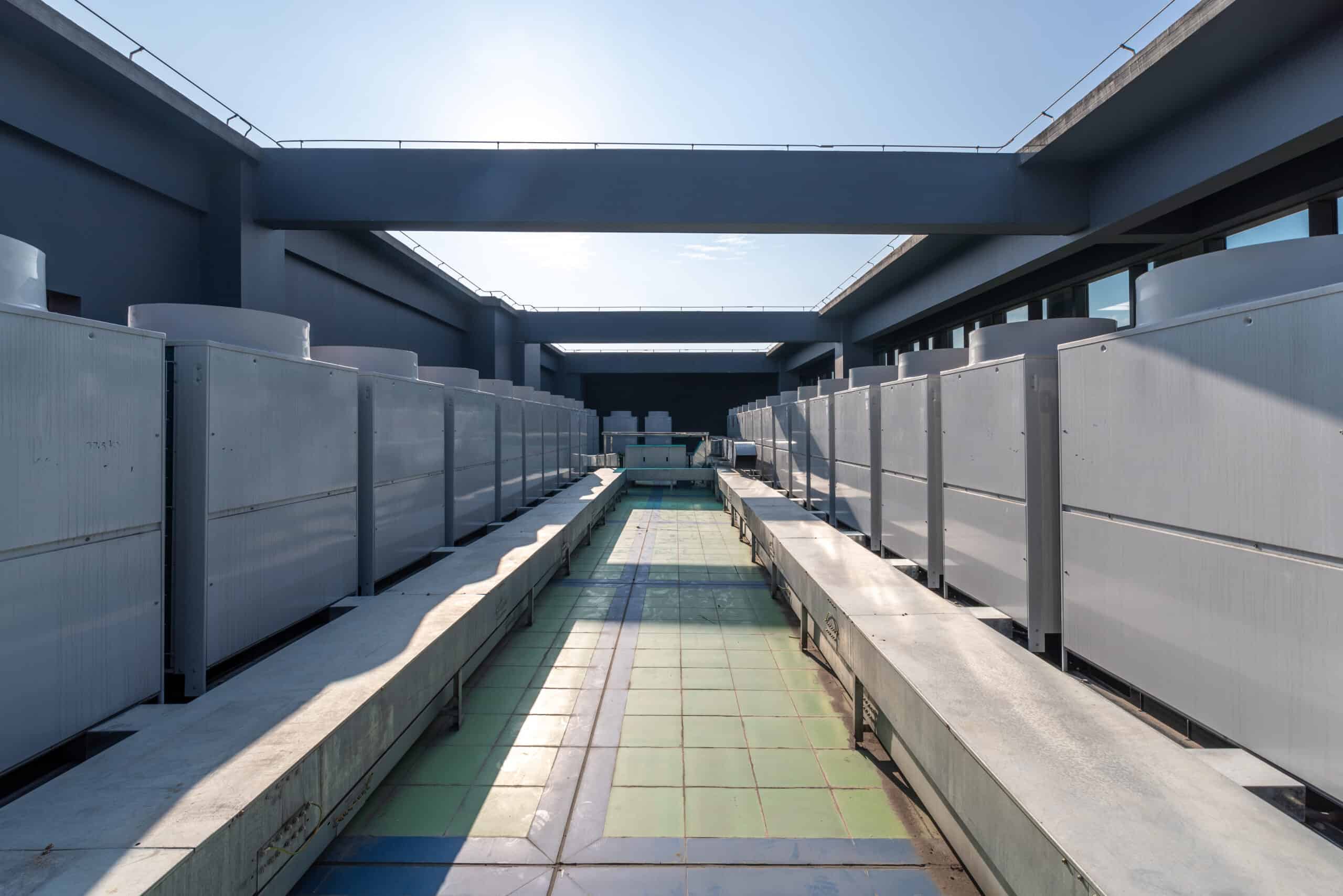 Rows of large rectangular HVAC units line both sides of a central tiled service walkway, flanked by metal platforms and overhead concrete beams on a rooftop courtyard under clear sky.