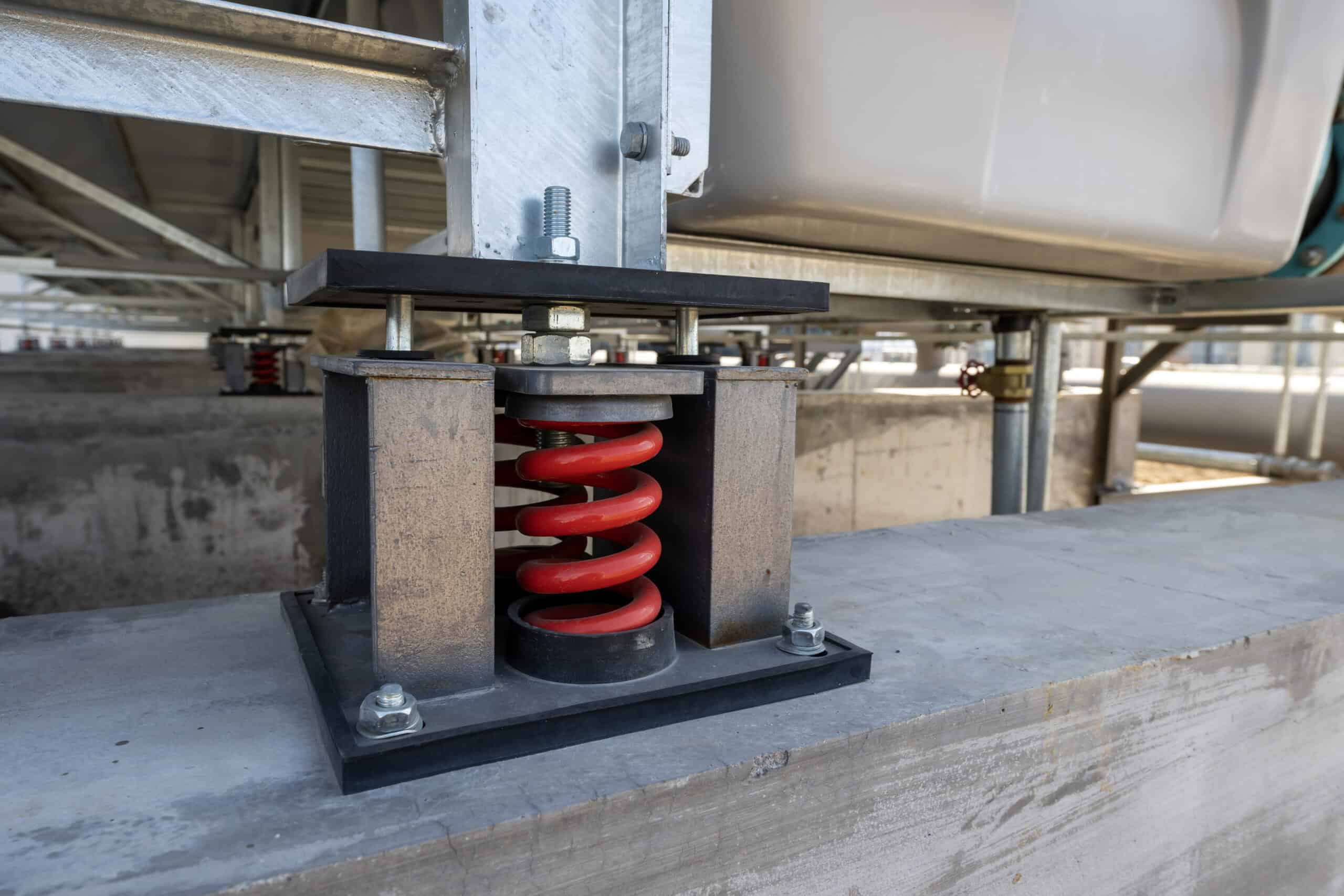 Red coil spring assembly supports a metal equipment frame, clamped between steel plates and bolted to a concrete base amid galvanized pipework and structural metal framing in an industrial rooftop/plant.
