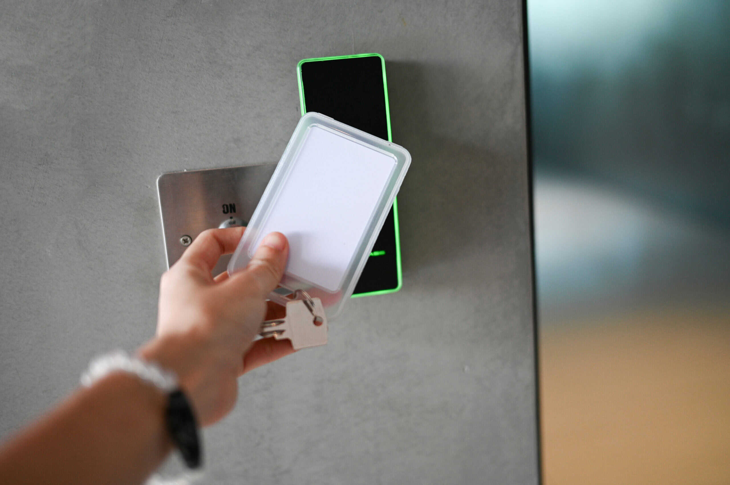 Keycard in clear plastic holder being tapped against a vertical black-and-green access reader on a gray metal door panel; nearby metal plate shows text: "ON".