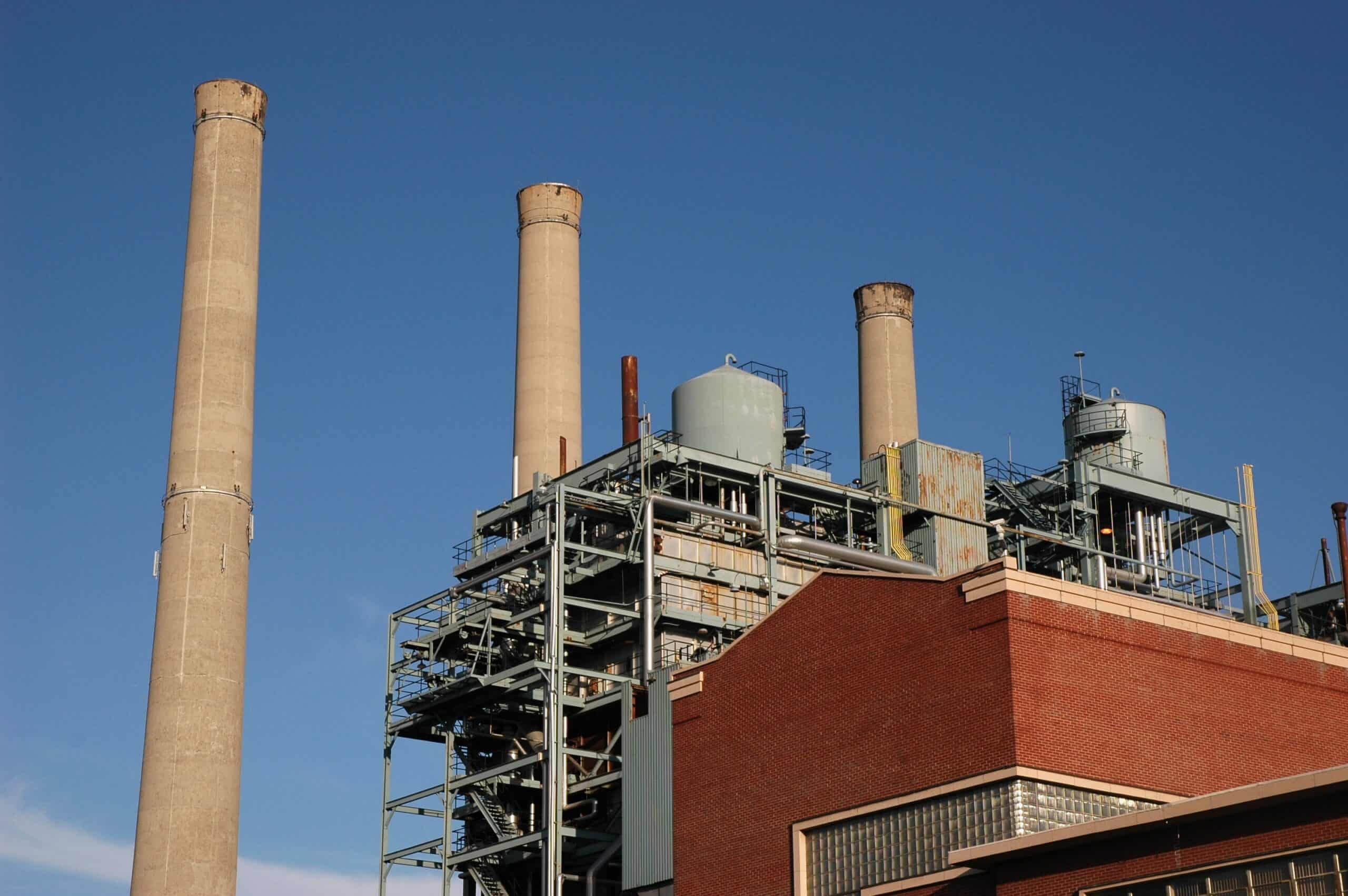 Four tall concrete smokestacks rise above a metal-framed industrial plant; piping, tanks and stairways crisscross atop a red-brick building under a clear blue sky.