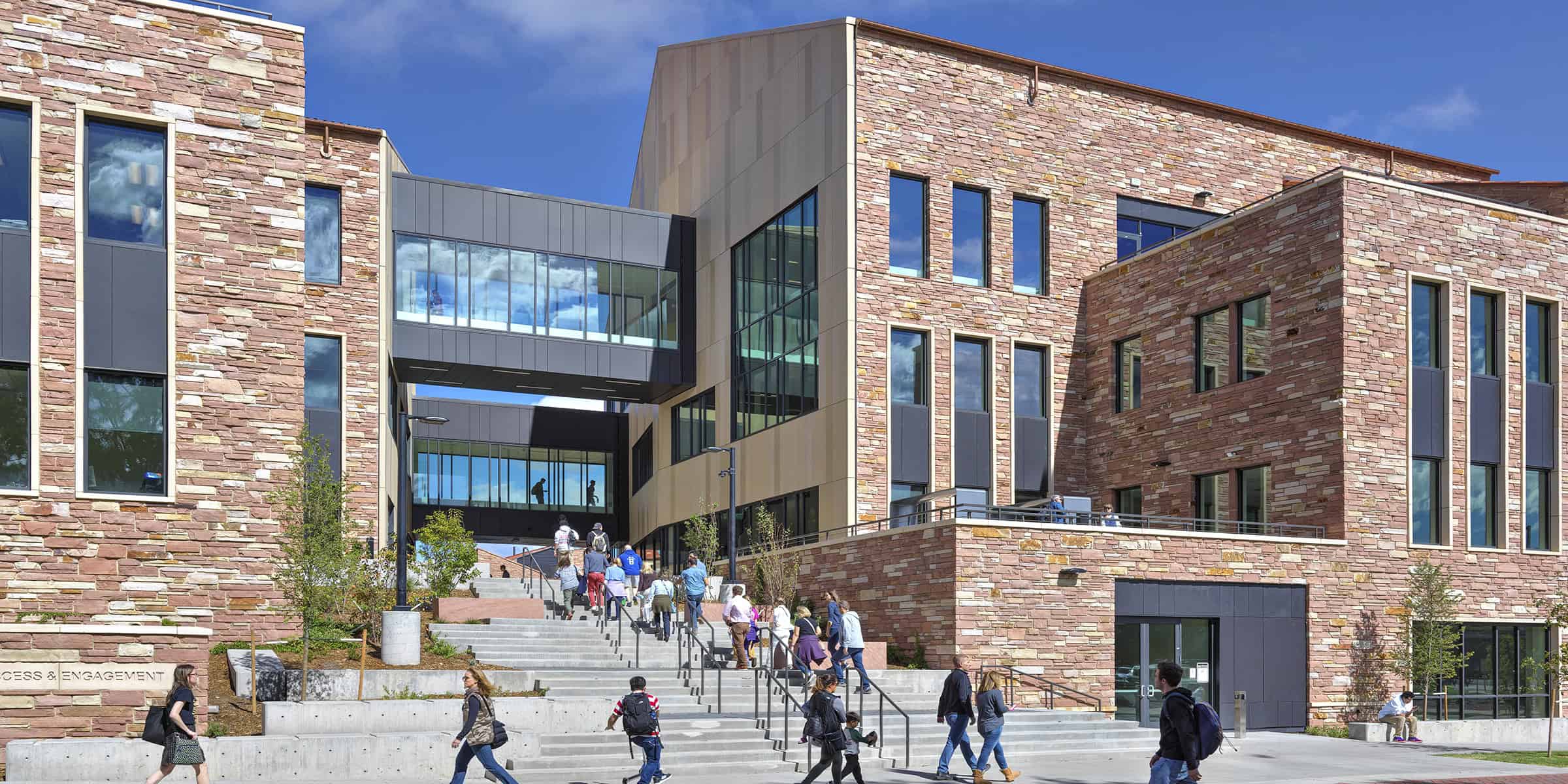 Stone-faced campus building; people ascend wide concrete steps toward glass skybridges, surrounded by planters and walkways under a bright blue sky. Text visible: "ACCESS & ENGAGEMENT".