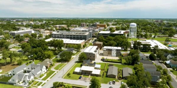 A white elevated water tower rises above a suburban neighborhood; houses and low-rise commercial buildings cluster below, with tree-lined streets, parking lots, lawns and a parking garage under cloudy sky.