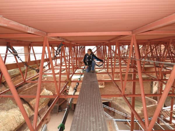 Person kneeling on a narrow elevated plank, clipped to safety cables and handling gear, amid red steel scaffolding under a corrugated roof above exposed rock and brick structures.
