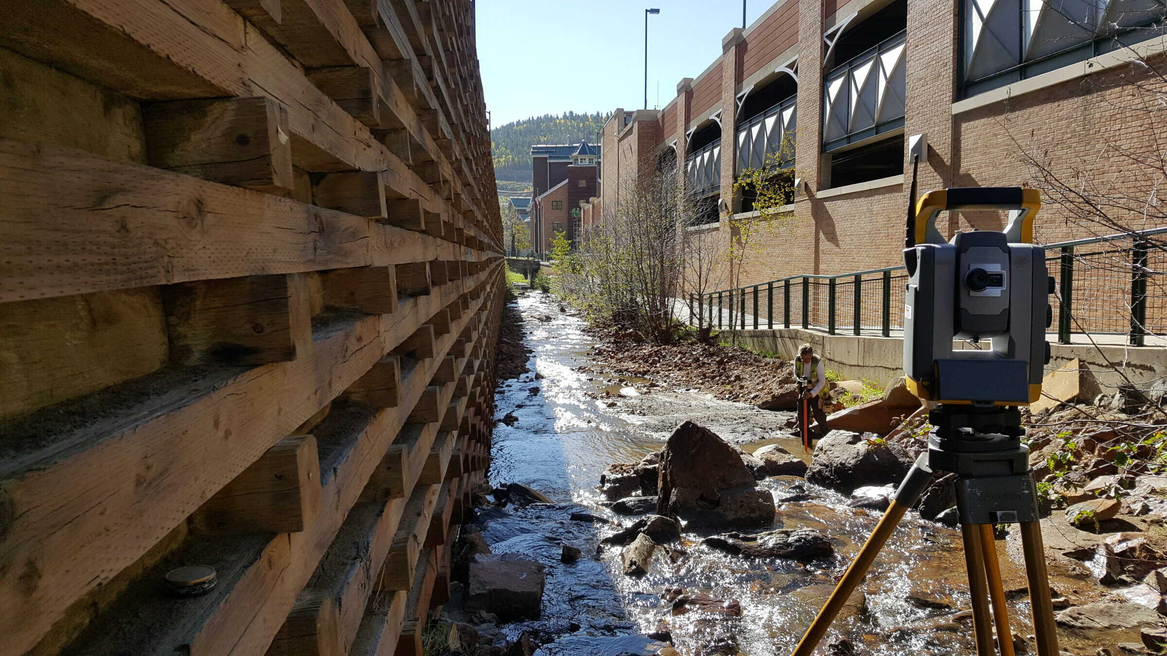 A surveying total station on a tripod records measurements while a worker holds a rod in a shallow rocky creek between a wooden retaining wall and adjacent brick buildings.