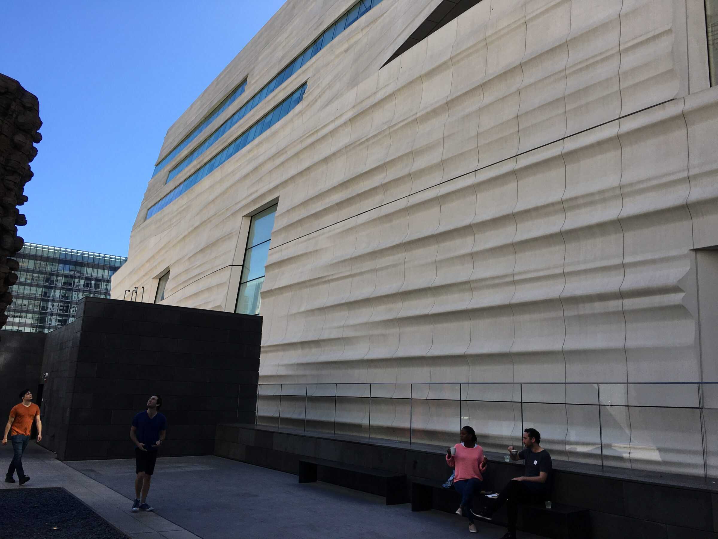Large sculpted white building facade with horizontal wave-like grooves; two people sit on a bench drinking, while two others walk and look up in a courtyard beneath blue sky.