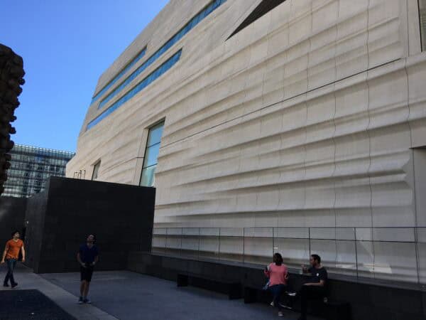 Large sculpted white building facade with horizontal wave-like grooves; two people sit on a bench drinking, while two others walk and look up in a courtyard beneath blue sky.