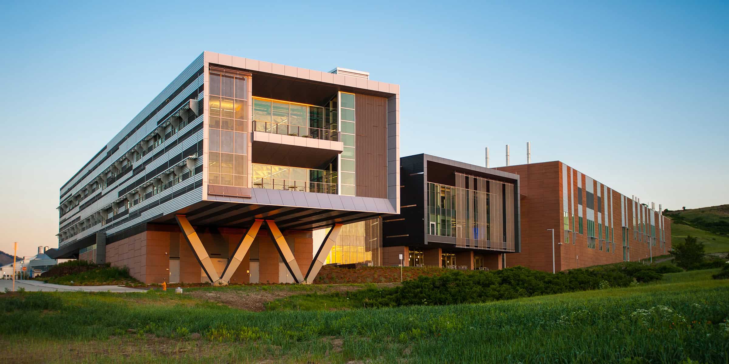 A modern multi-story research building cantilevers on V-shaped supports, its glass-walled upper floors glowing with interior light against a clear evening sky, set above grassy hills.