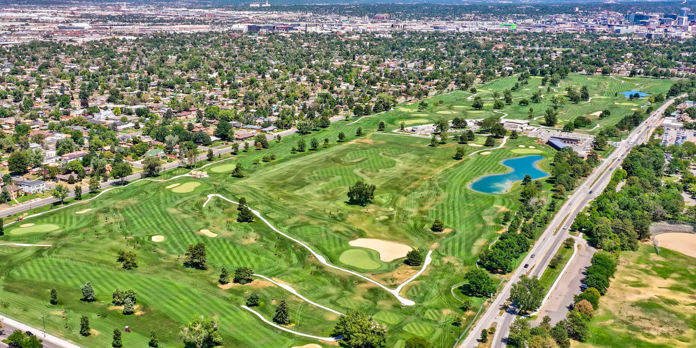 Golf course stretches across the foreground with striped fairways, sand bunkers and blue ponds; bordered by a residential neighborhood, roads and a distant city skyline.