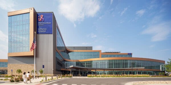 Glass-clad hospital building curves forward with a covered entrance; an American flag flies as two people walk across a landscaped campus. Text: "ih Intermountain Health Lutheran Hospital"