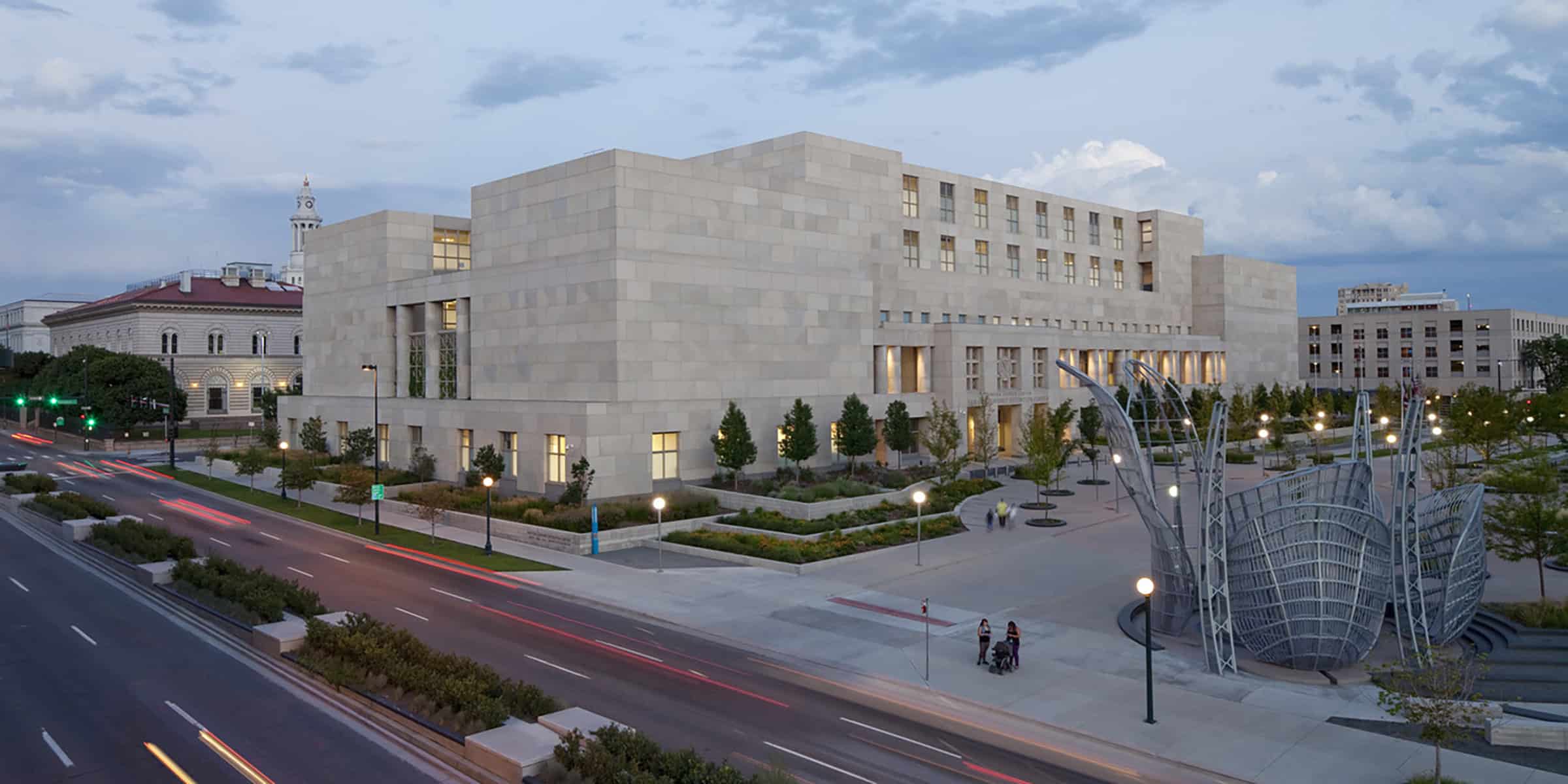 Large limestone civic building stands beside a plaza, windows glowing as people walk; a metal sculpture anchors the plaza while vehicle light trails streak along the adjacent boulevard at dusk.