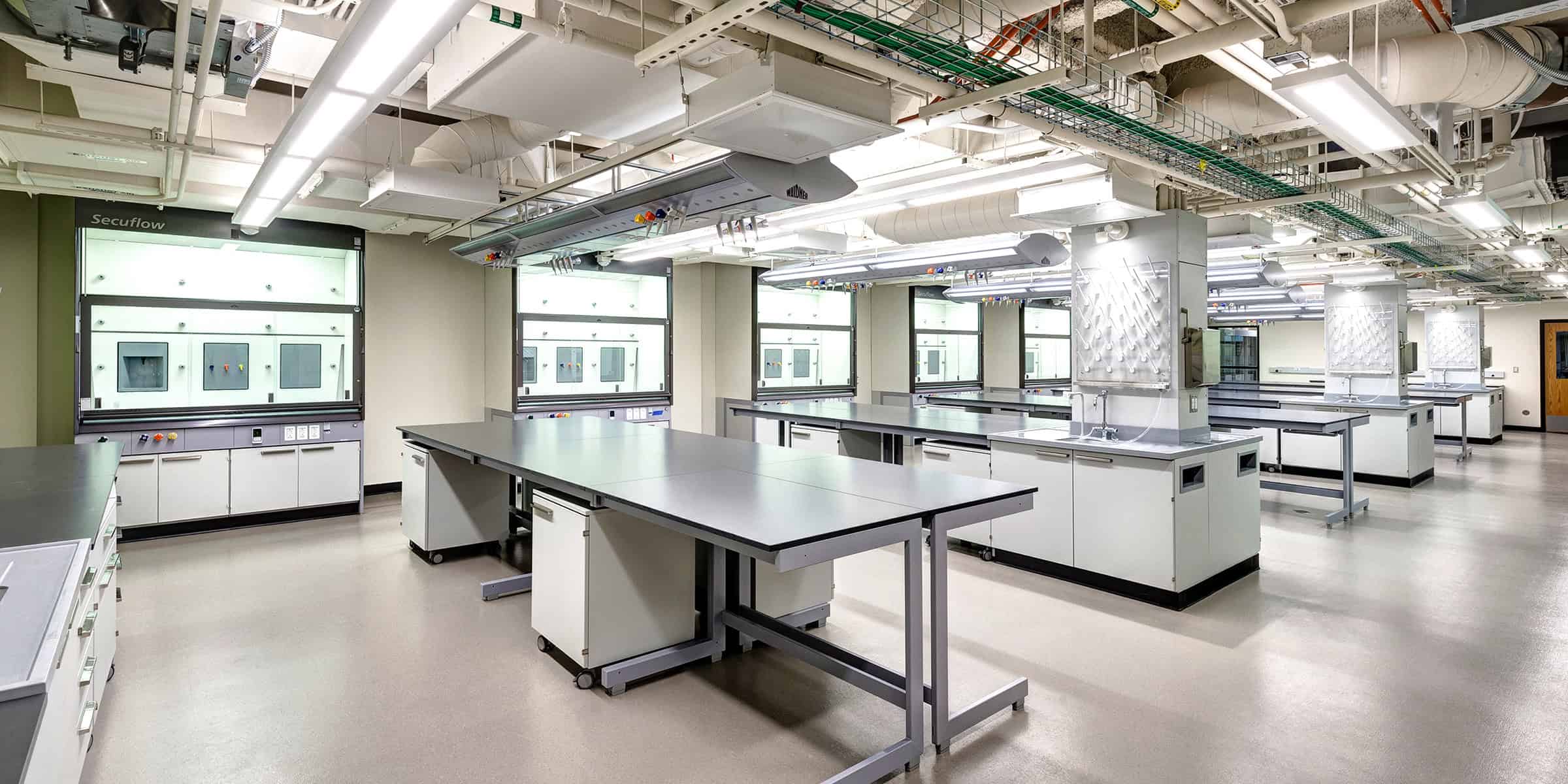 Laboratory workbenches stand empty amid illuminated fume hoods and overhead service units in a spacious, modern chemistry lab with sinks and storage. Text: "Secuflow" (on fume hood), "WALDNER" (on overhead units).