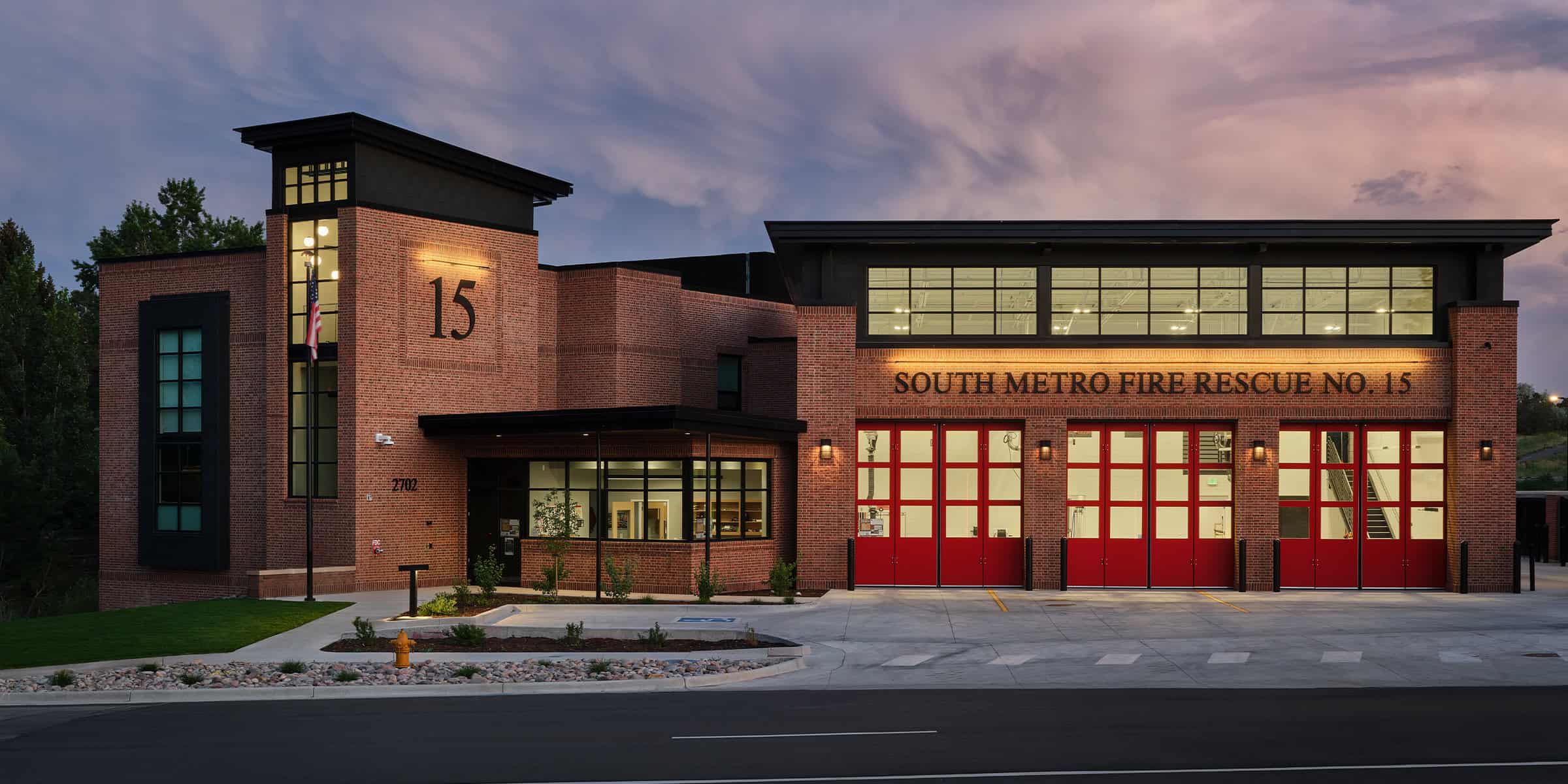 Brick fire station sits illuminated at dusk, displaying red garage doors and an American flag, fronted by a paved driveway and landscaped curb in a quiet suburban setting.

Text found in the image:
15
SOUTH METRO FIRE RESCUE NO. 15
2702