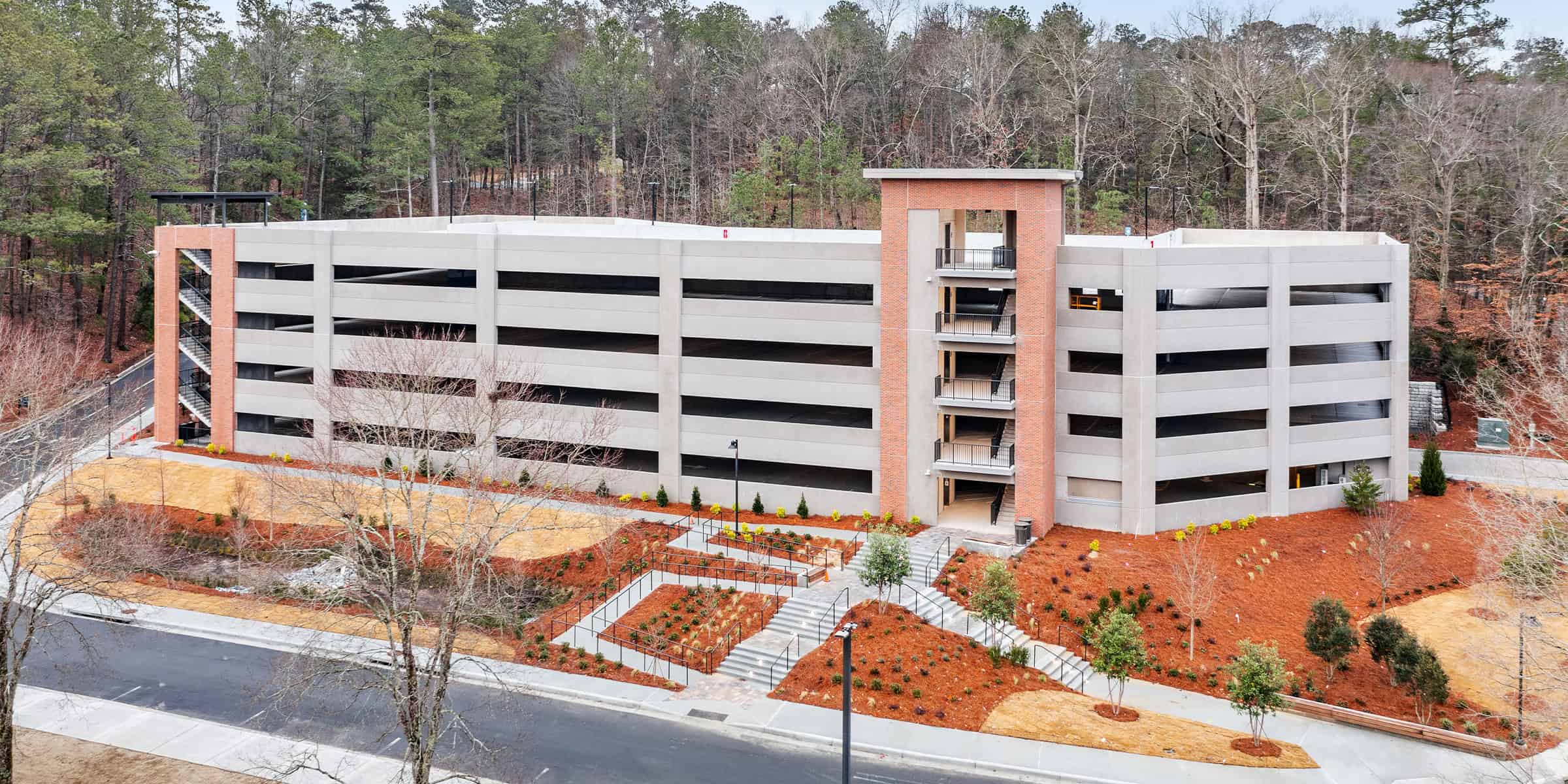 Multi-level concrete parking garage rises with exposed stair towers and open horizontal bays, flanked by brick cores and landscaped terraced steps, adjacent to a road and wooded hillside.