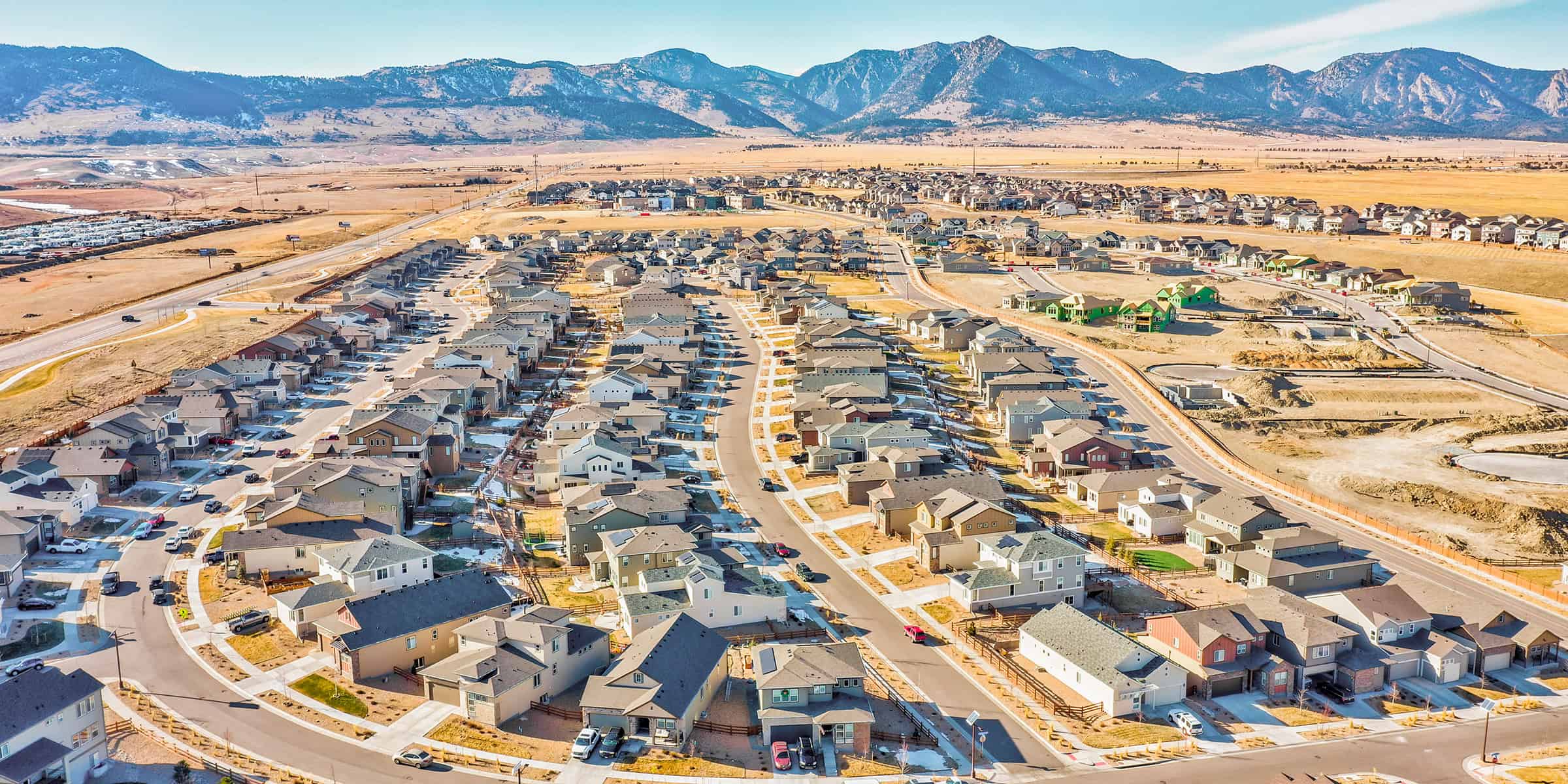 Suburban neighborhood: rows of houses line winding streets, some new homes under construction; development sits amid dry fields and lots, with rugged mountains beyond under a clear sky.