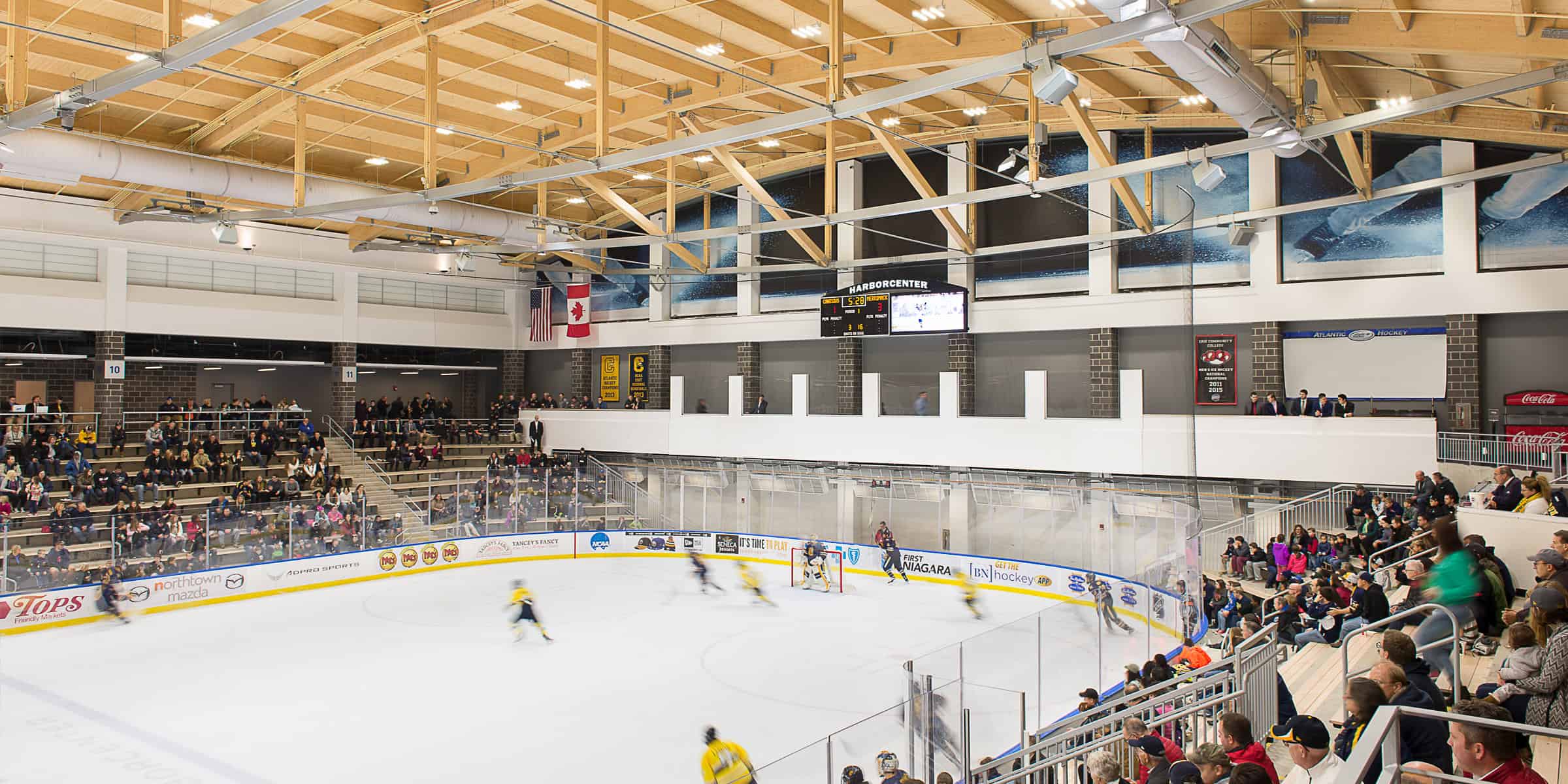 Hockey players skate toward a net while a goalie defends, spectators fill bleachers under exposed wooden rafters and a scoreboard reads HARBORCENTER.

Transcribed text: HARBORCENTER; TOPS Friendly Markets; northtown mazda; FIRST NIAGARA; GET THE hockey APP; IT'S TIME TO PLAY; ATLANTIC HOCKEY; Coca-Cola