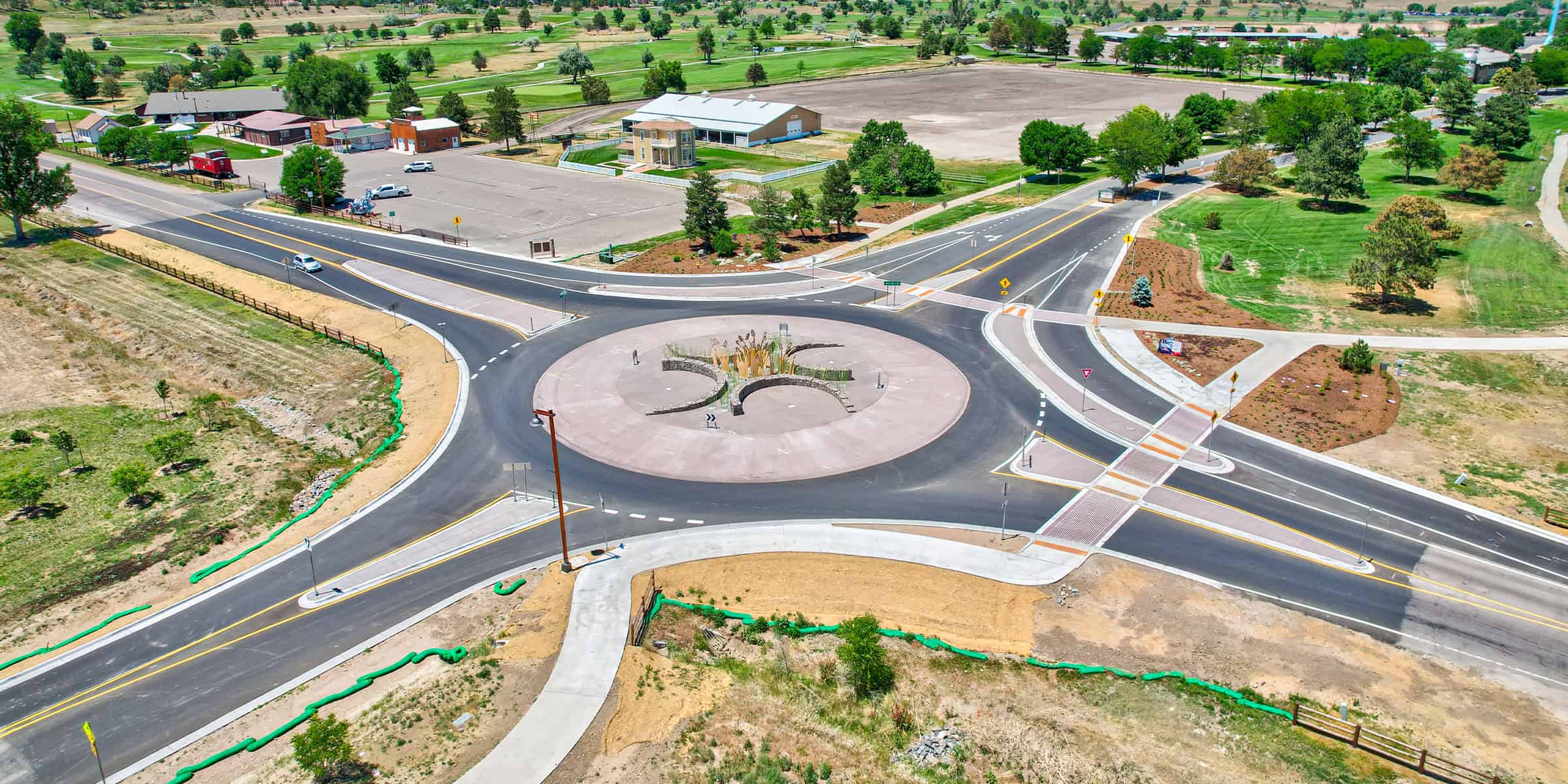 A large circular roundabout channels traffic around a landscaped central island with sculptural planting, connecting four asphalt roadways amid grassy fields, sidewalks, a parking lot, and scattered buildings.