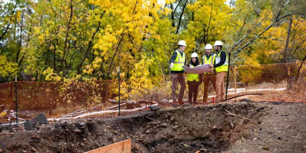 Group of four construction workers in hard hats and high-visibility vests consult blueprints while standing behind orange safety fencing beside an excavation pit, surrounded by yellow autumn trees.