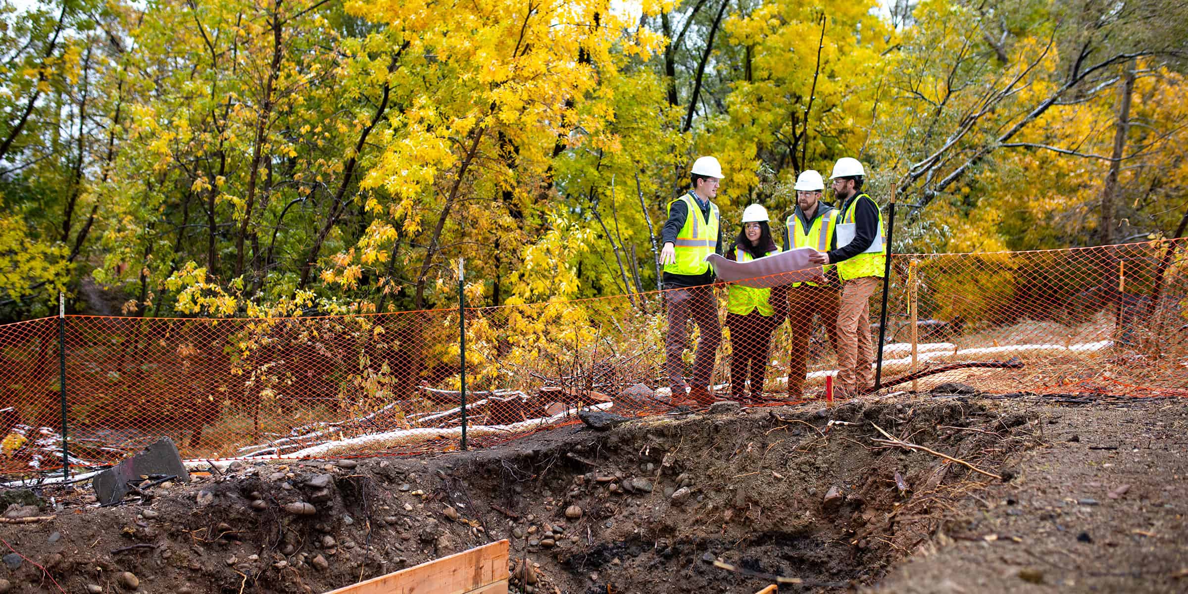 Group of four construction workers in hard hats and high-visibility vests consult blueprints while standing behind orange safety fencing beside an excavation pit, surrounded by yellow autumn trees.