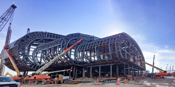 Curved steel-framed building shell rising, surrounded by cranes and boom lifts reaching into its structure on a muddy construction site beneath a clear blue sky.

Text in image: "JLG ULTRA BOOM", "United Rentals", "JLG"