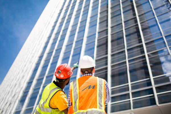 Two construction workers in hard hats and reflective vests point and inspect a tall glass office tower rising against a clear blue sky. Text on vest: "m".