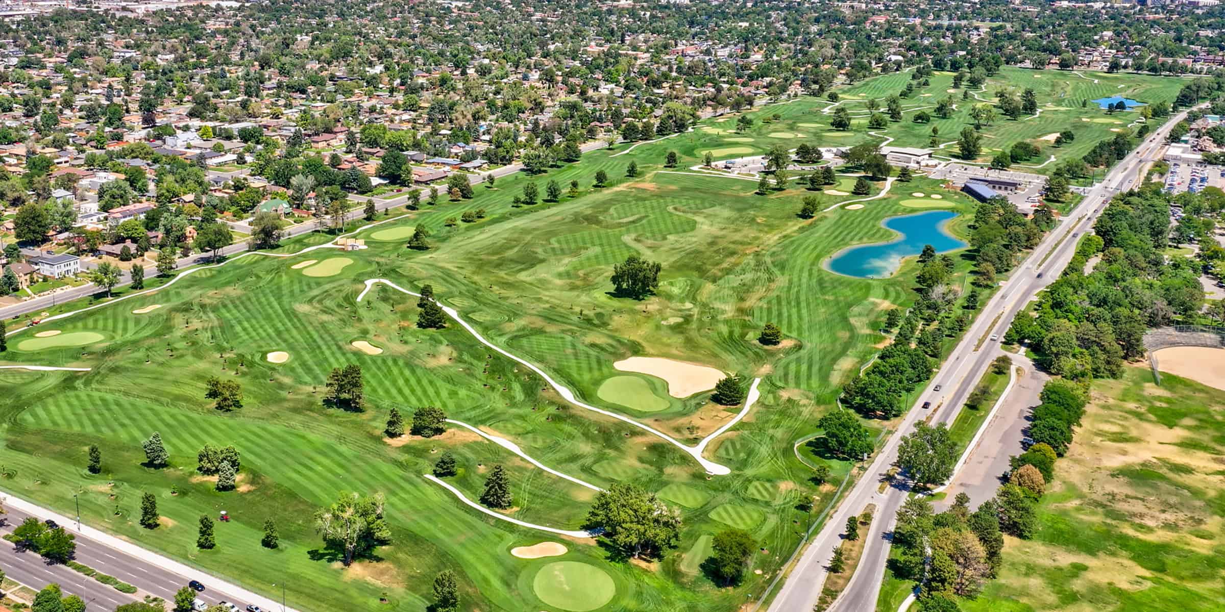 A sprawling golf course stretches with striped fairways, sand bunkers and blue water hazards, bordered by a suburban neighborhood and parallel roads with scattered trees and small ponds.