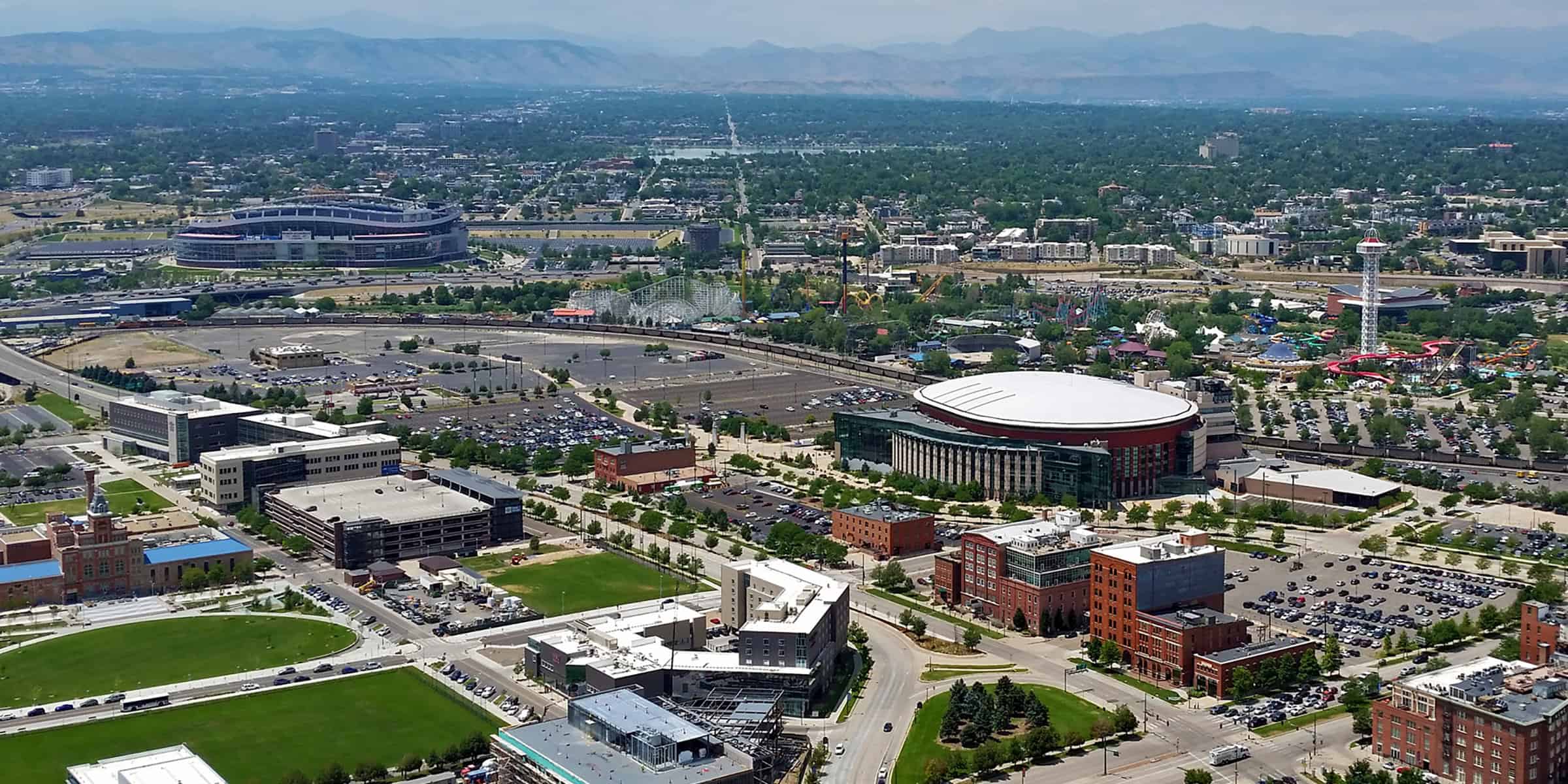 A domed arena sits amid parking lots and mid-rise buildings; a larger open-air stadium and amusement park lie nearby, all within a sprawling cityscape with distant mountains.