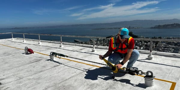 A construction worker crouches on a rooftop, tightening a yellow safety line and chain anchor with a tool; white roof, guardrail, city buildings and bay visible under a blue sky.